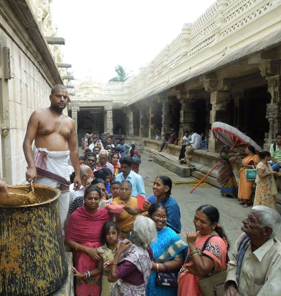 A vaidika standing over a great vessel of rice ladling it out to the queue pressed along the temple corridor beneath him — women, children and old men holding their plates up, the carved stone colonnade running away behind them