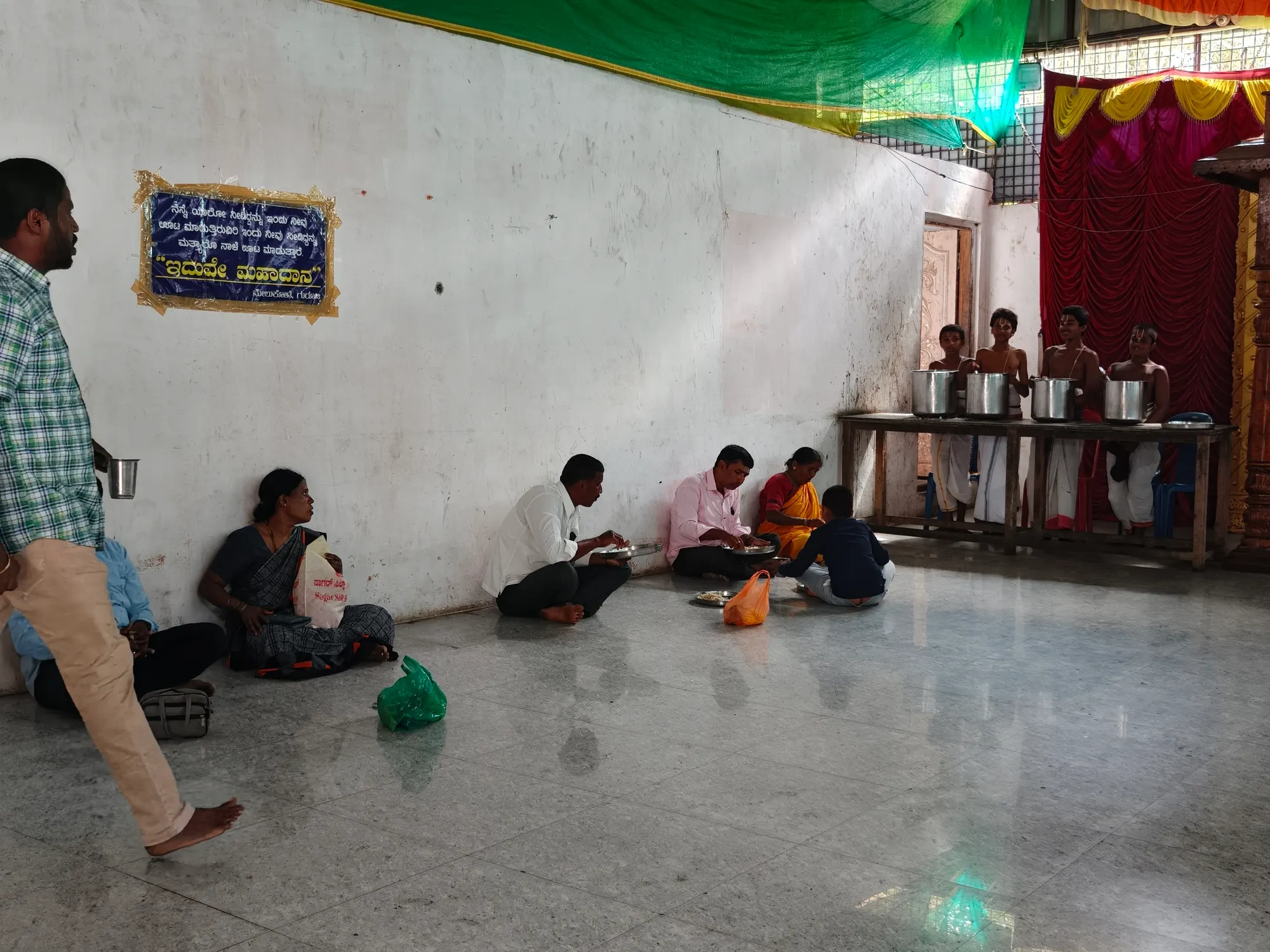 Pilgrims seated along the wall of the annadana hall eating from steel plates while gurukulam students stand ready behind the serving vessels; the Kannada plaque on the wall reads annadana mahadana