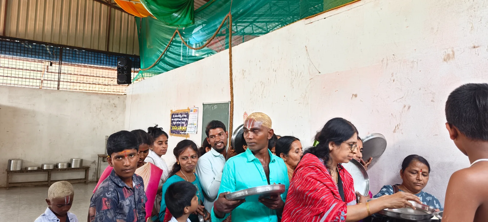 Pilgrims carrying filled steel plates back through the crowd of the annadana hall, children among them, the stacked vessels ranged along the wall behind