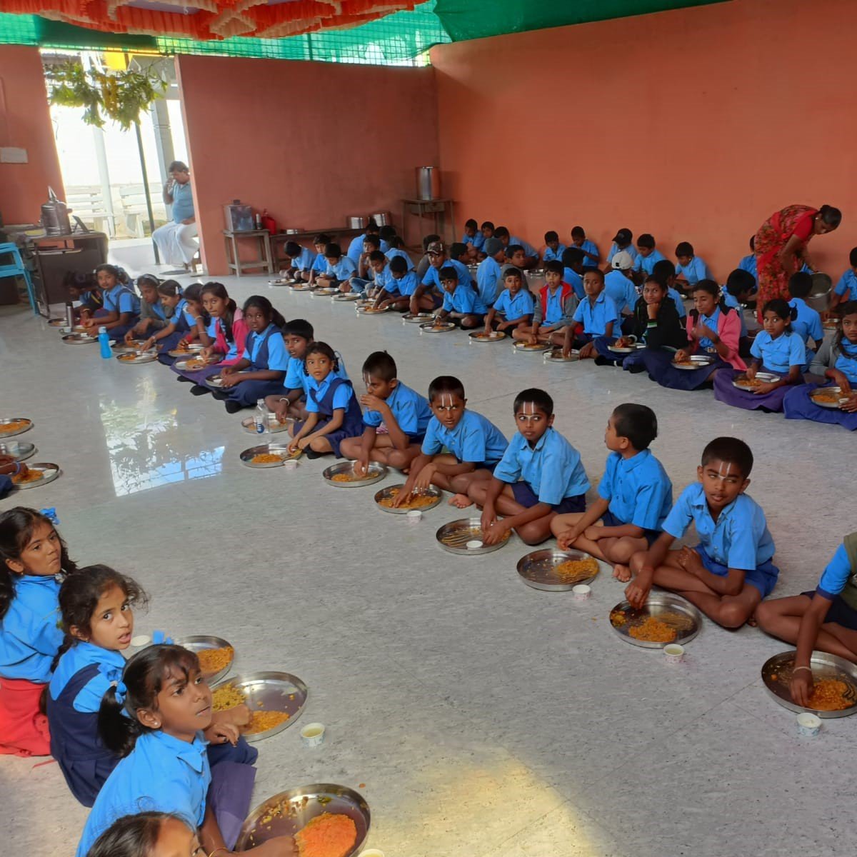 Village schoolchildren in blue uniforms seated in long rows on the floor, eating a hot meal from steel plates