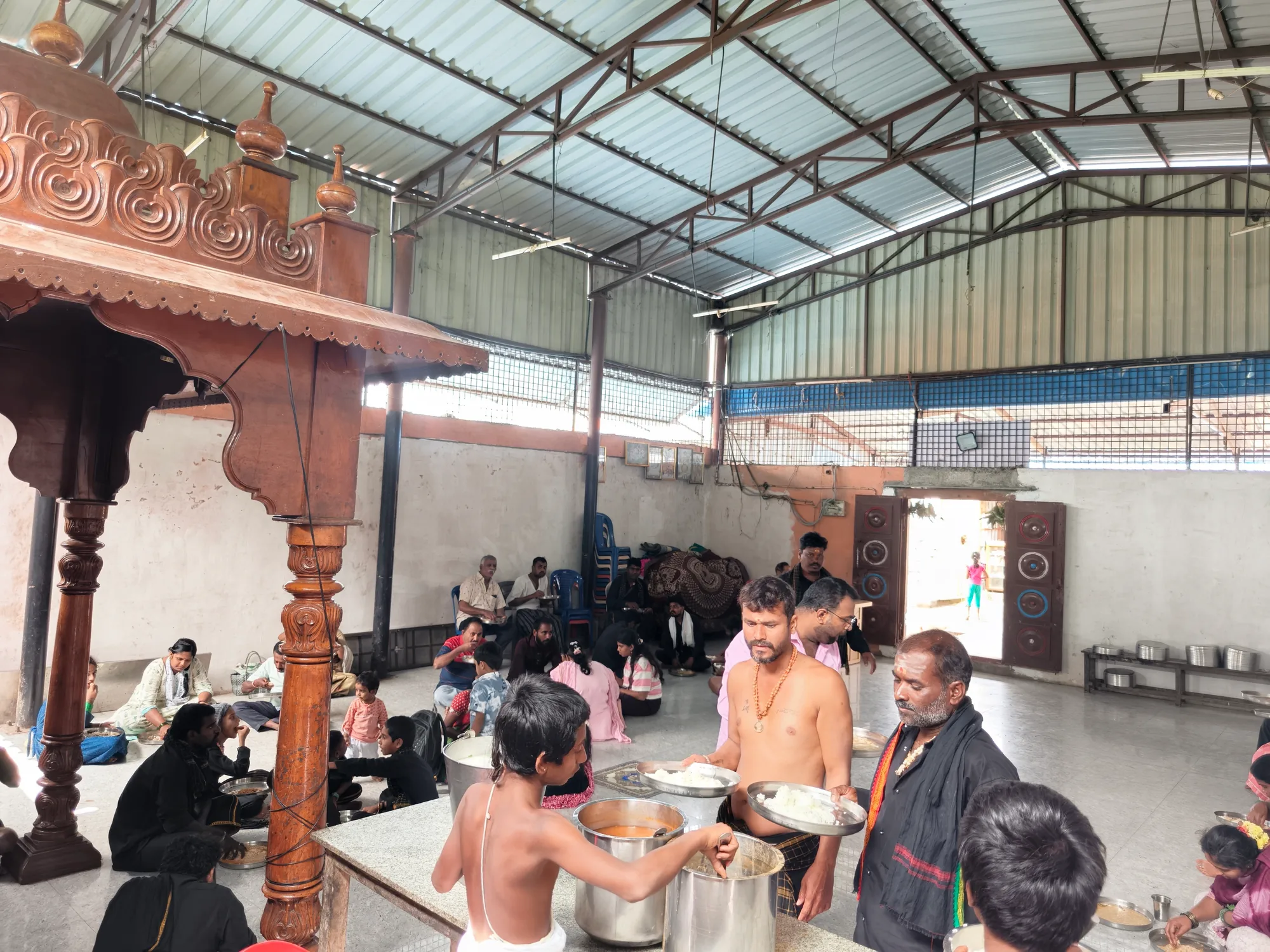The annadana hall under its steel roof at midday, a boy in a dhoti and a server carrying steel vessels of rice between the rows, families already seated and eating along the far wall