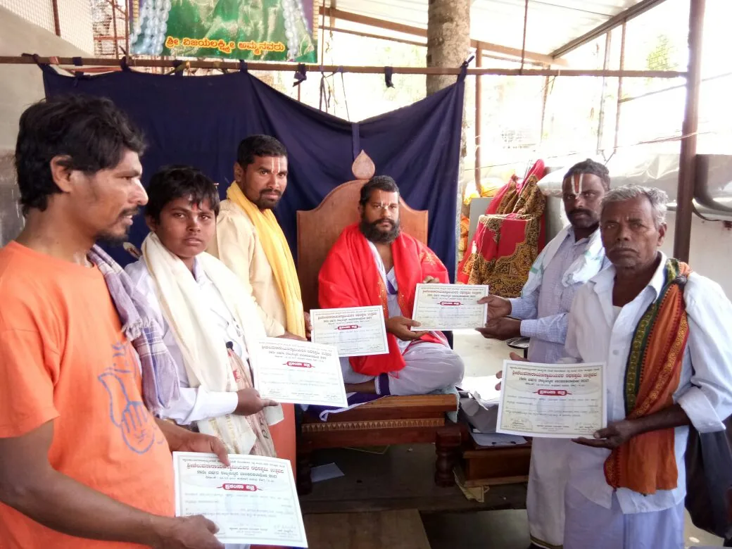 Men of the village standing in a row each holding a printed certificate, the vaidika who gave them seated behind on a wooden seat