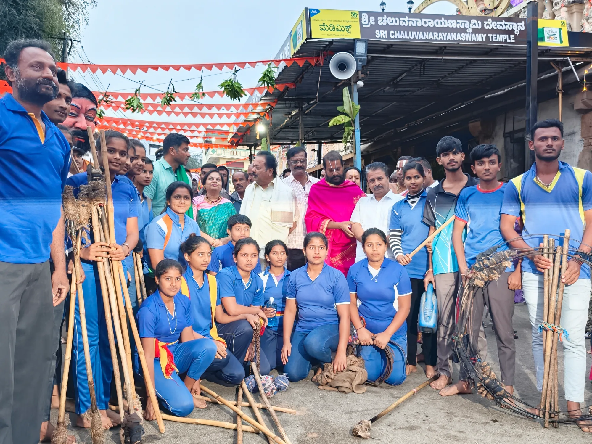 Schoolchildren and volunteers with brooms and rakes gathered at the temple gate for a cleaning drive, the temple's board above them