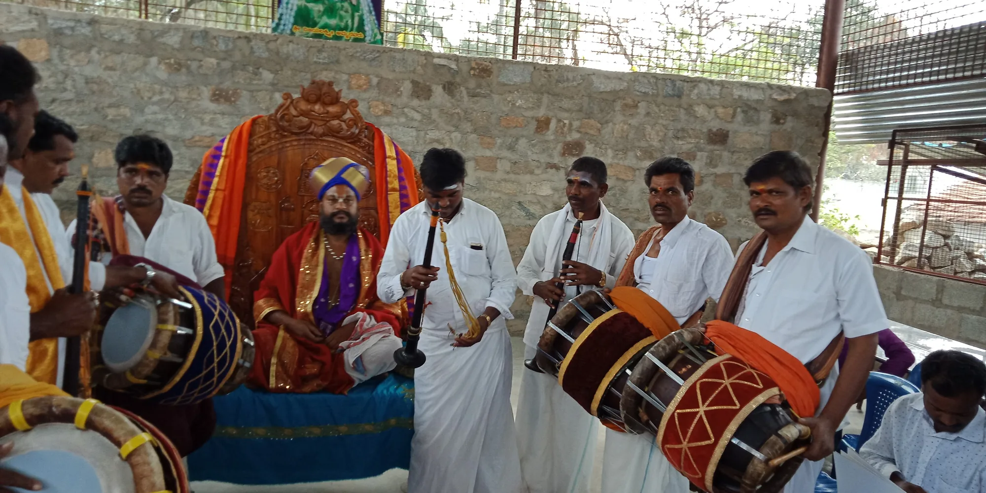 A line of nadaswara and drum players performing in front of Guruji, who is seated in a turban and rose silk on a carved seat