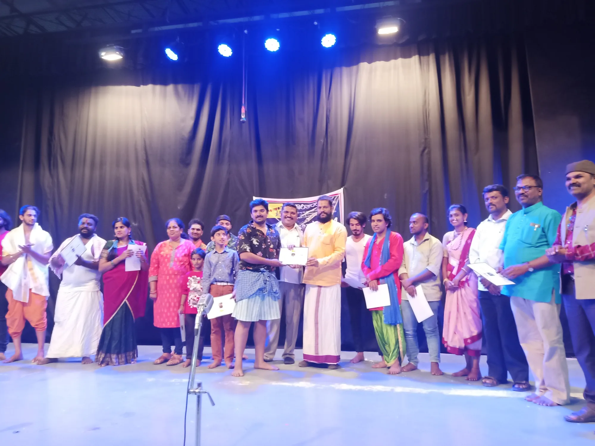 The cast of a folk drama in costume lined across the stage after the performance while one of them is presented with a memento