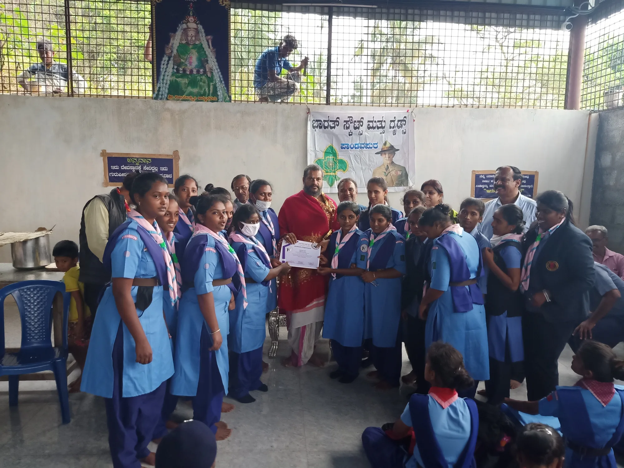 A troop of Guides in uniform crowded around a vaidika in red holding a framed certificate, the movement's banner on the wall behind