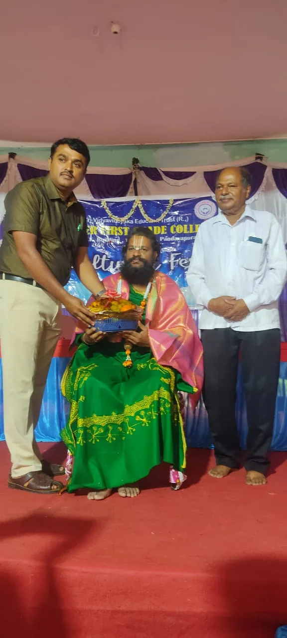 A vaidika in green seated on a college stage taking a shawl and an offering from two members of the staff, the event's banner behind