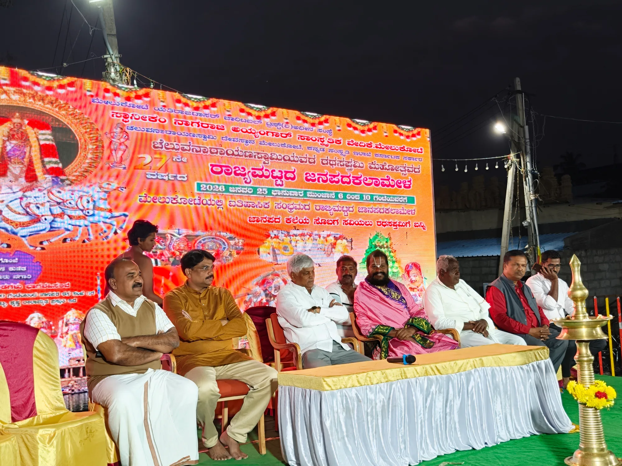 The guests of a folk-arts festival seated across a stage at night under a long printed banner, a brass lamp standing lit at the right