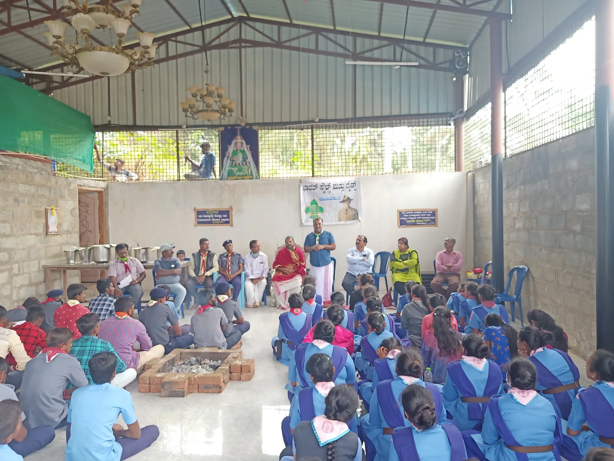 A hall of schoolchildren in uniform seated on the floor before a line of teachers and guests, a brick homa kunda in the middle of the floor