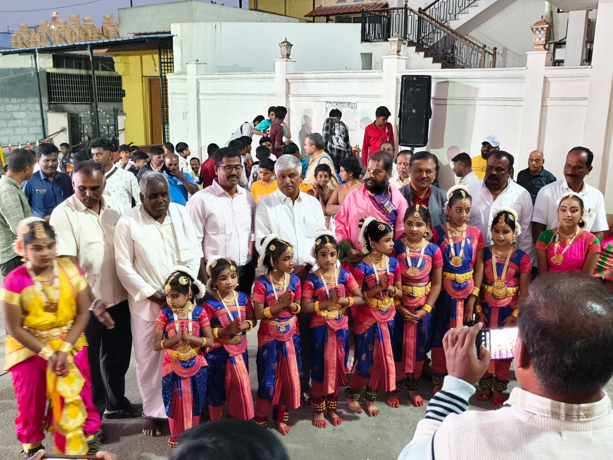 A line of small girls in dance costume and gold jewellery standing with the organisers outside the hall before their performance