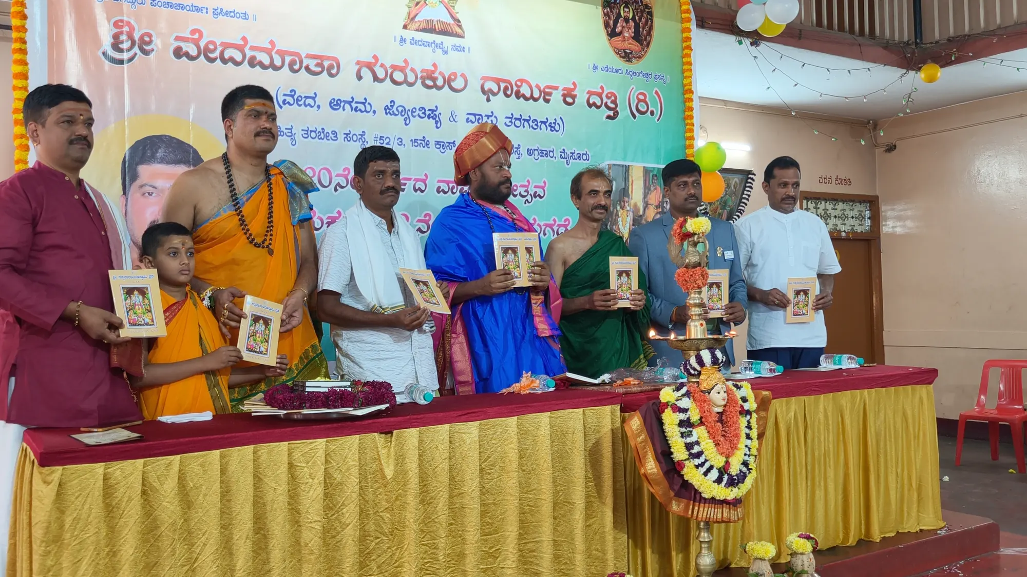 A book being released on the gurukulam's stage, each of the guests holding a copy up beside the lit lamp