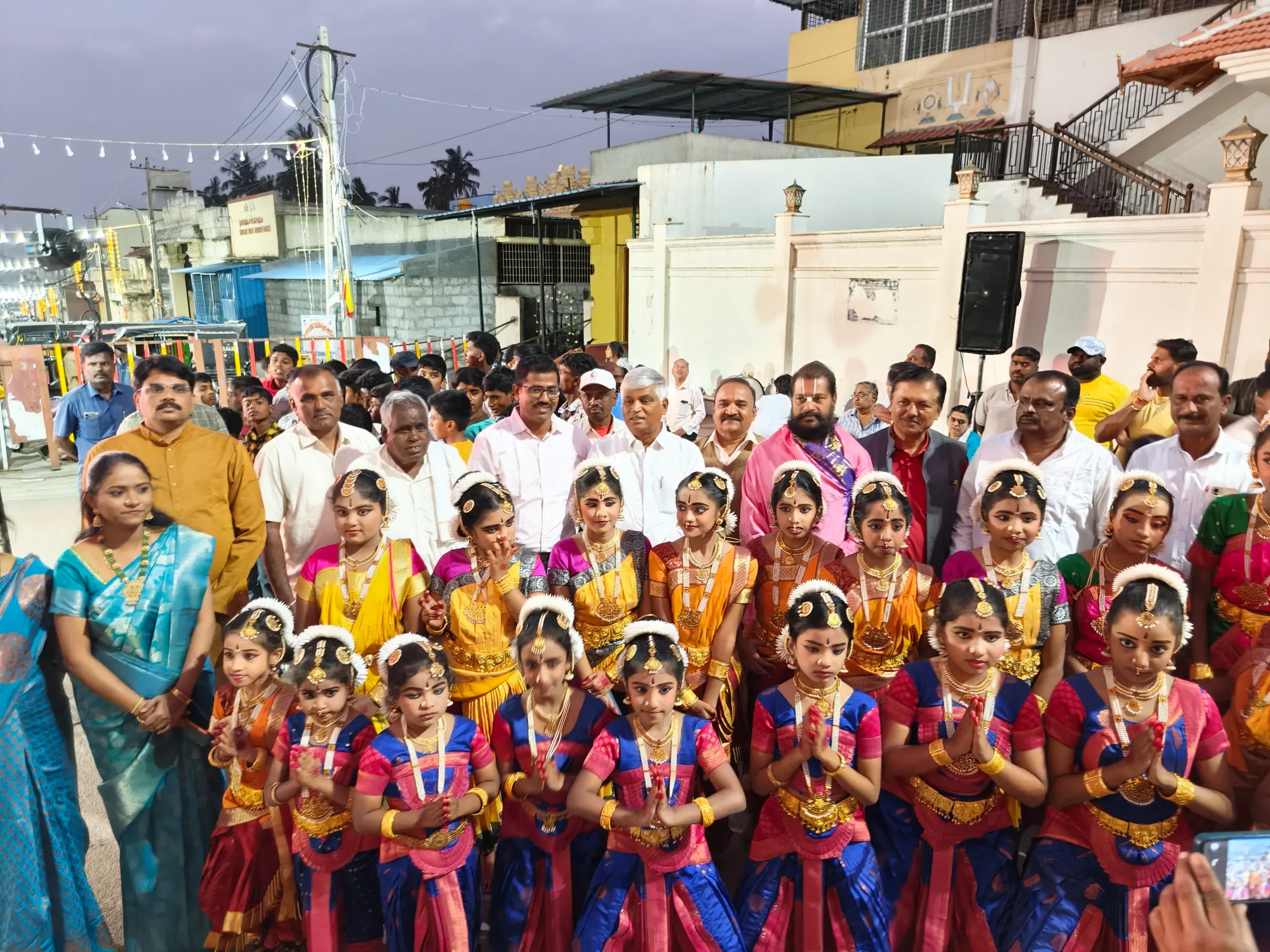 Young Bharatanatyam dancers in full costume standing with palms joined in the temple street at dusk, the adults of the programme behind them