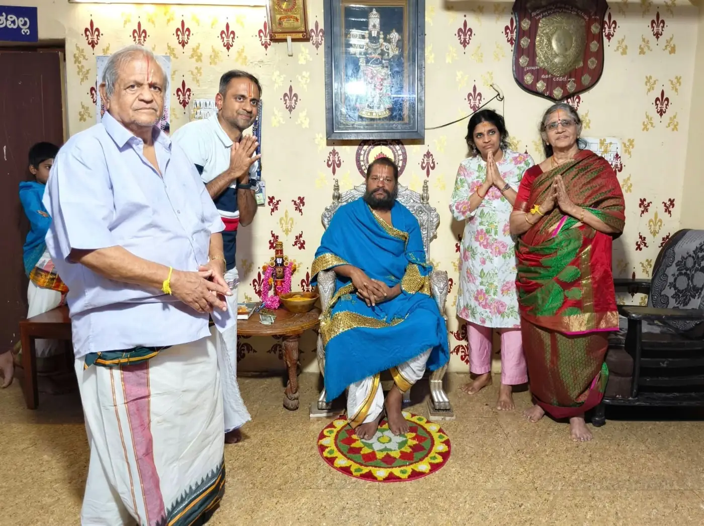 The elders of a family standing with palms joined on either side of Guruji, seated in a blue and gold shawl with a lamp and a small shrine at his side