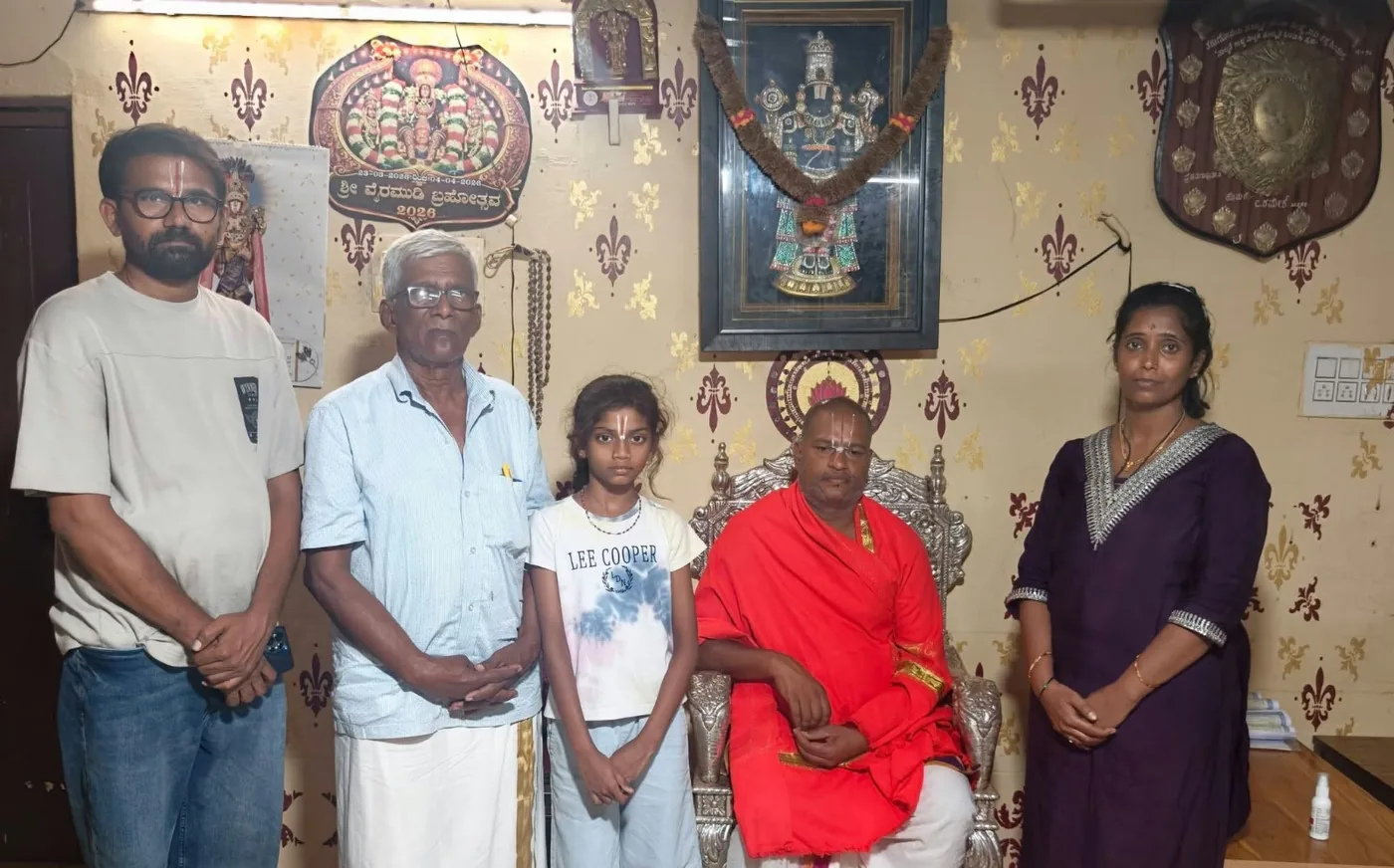 A man, a woman, an elderly father and a girl standing around Guruji seated in a red shawl, the framed deity of the mandira on the wall behind them