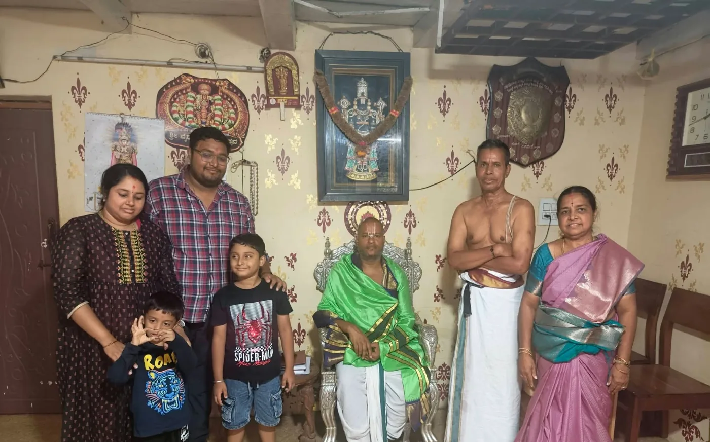 A family with two young sons standing beside Guruji, seated in a green shawl, a vaidika of the mandira standing at his other side