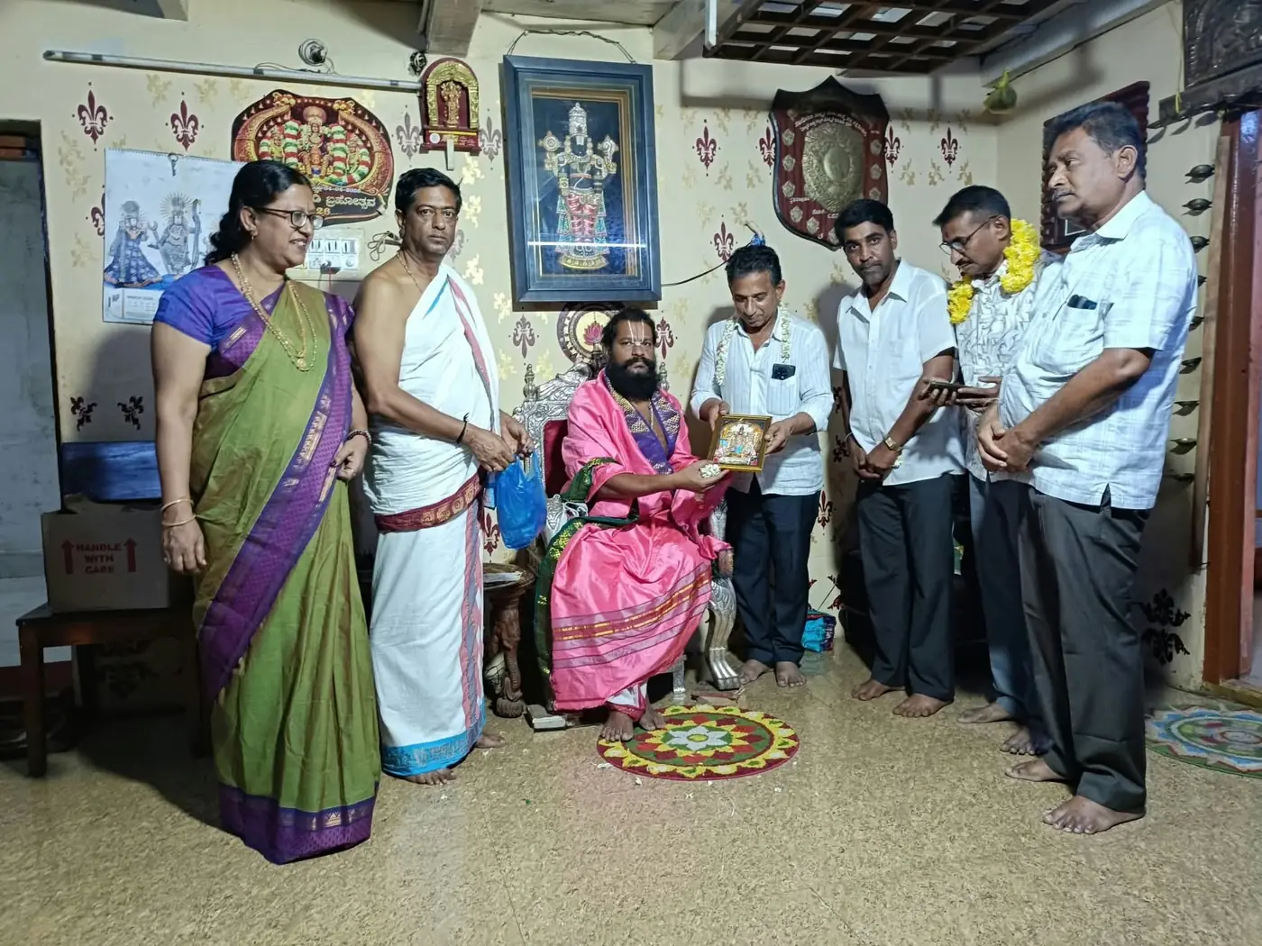 A framed picture of the deity handed across to a visitor by Guruji, seated in a rose shawl, with the visitor's family and a garlanded man standing around them