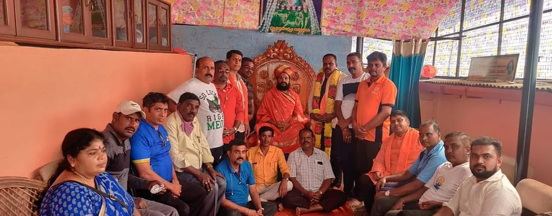 A large party seated on the floor in rows around Guruji's chair, filling the width of the mandira room, everyone turned to the camera