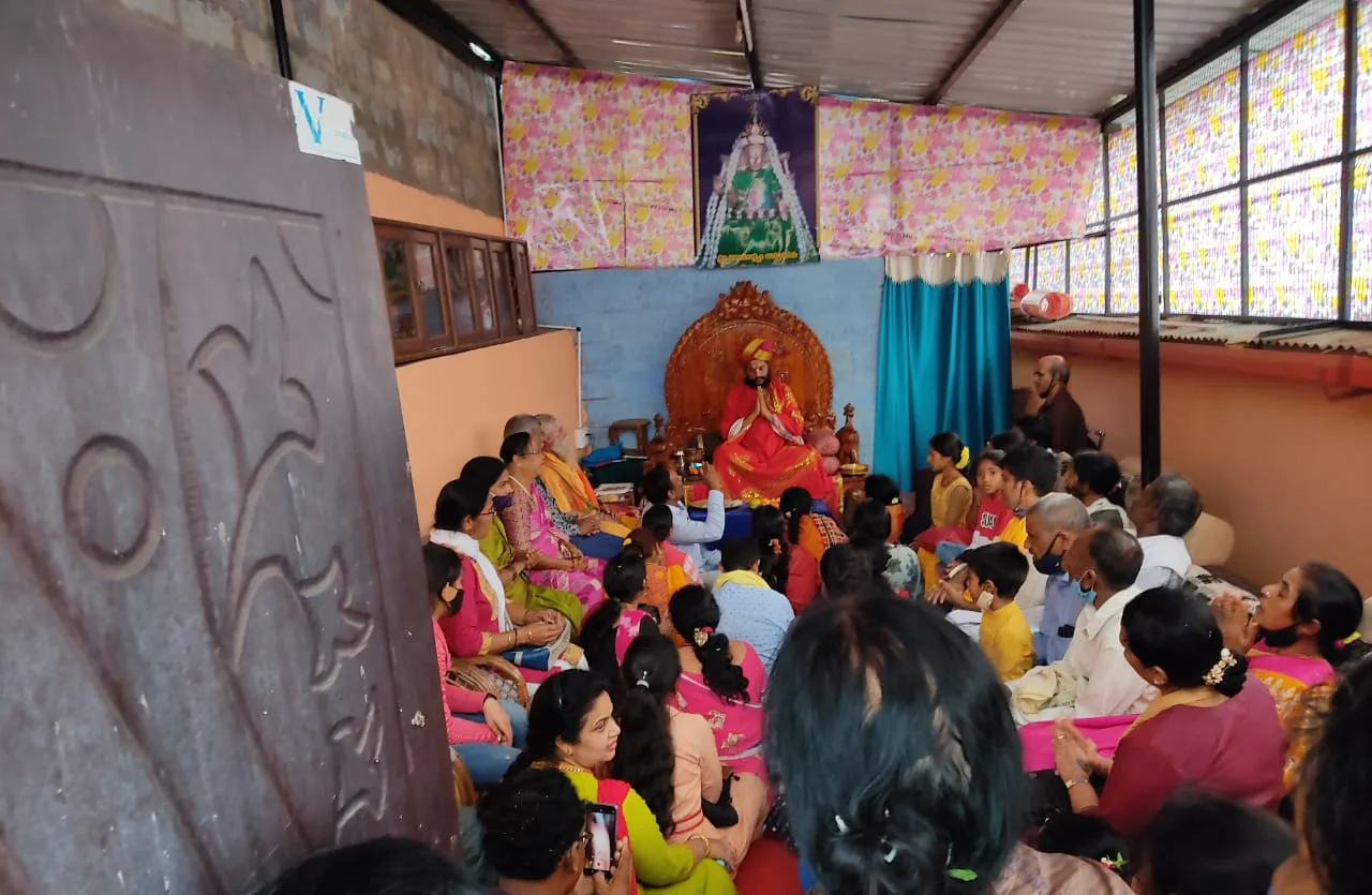 The mandira hall packed wall to wall with seated devotees, families on the floor and children among them, Guruji on the carved throne at the far end beneath a painted backdrop