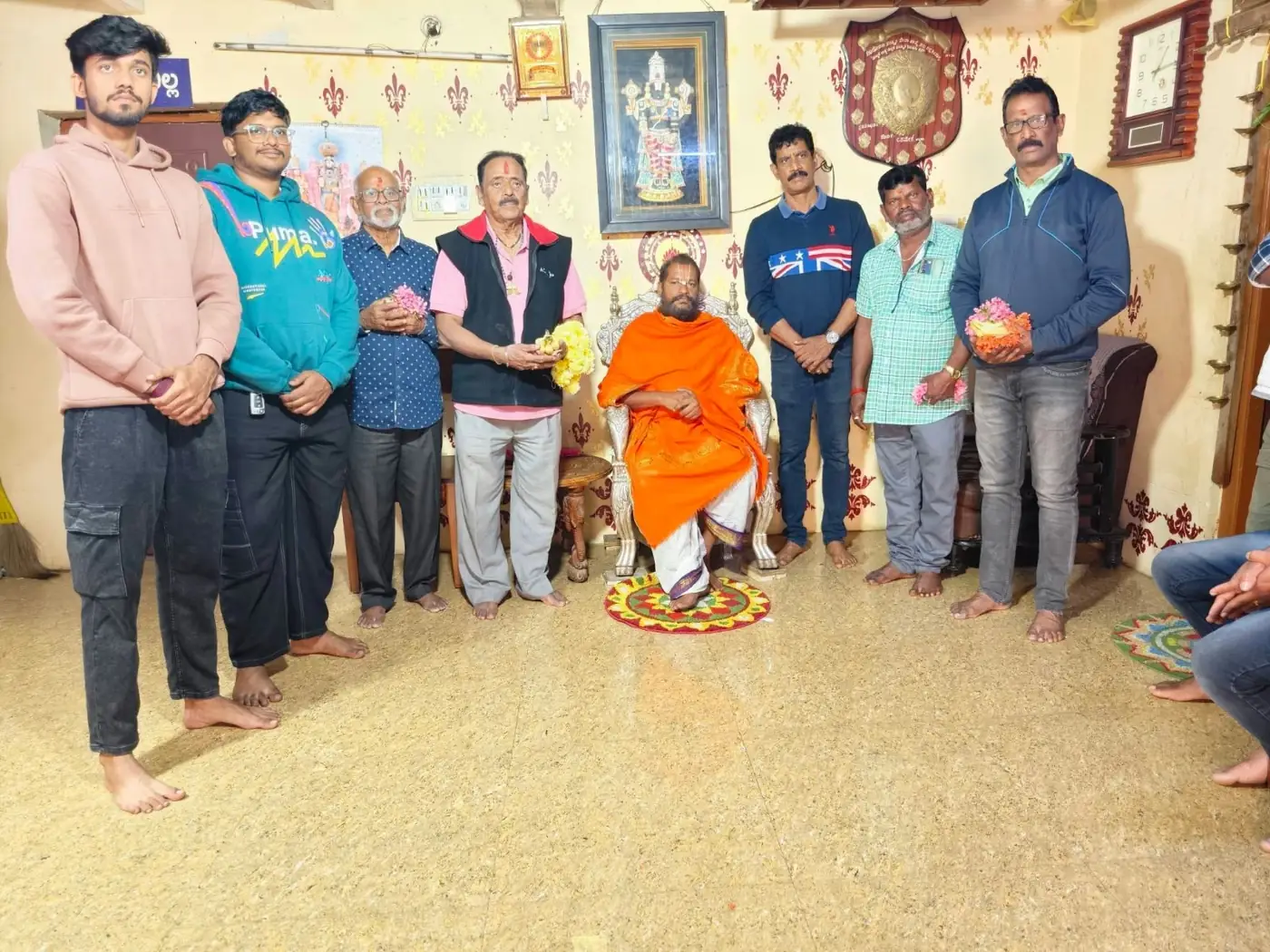 Eight men standing along both walls of the mandira room, two of them holding offering plates, Guruji seated in an orange shawl at the centre