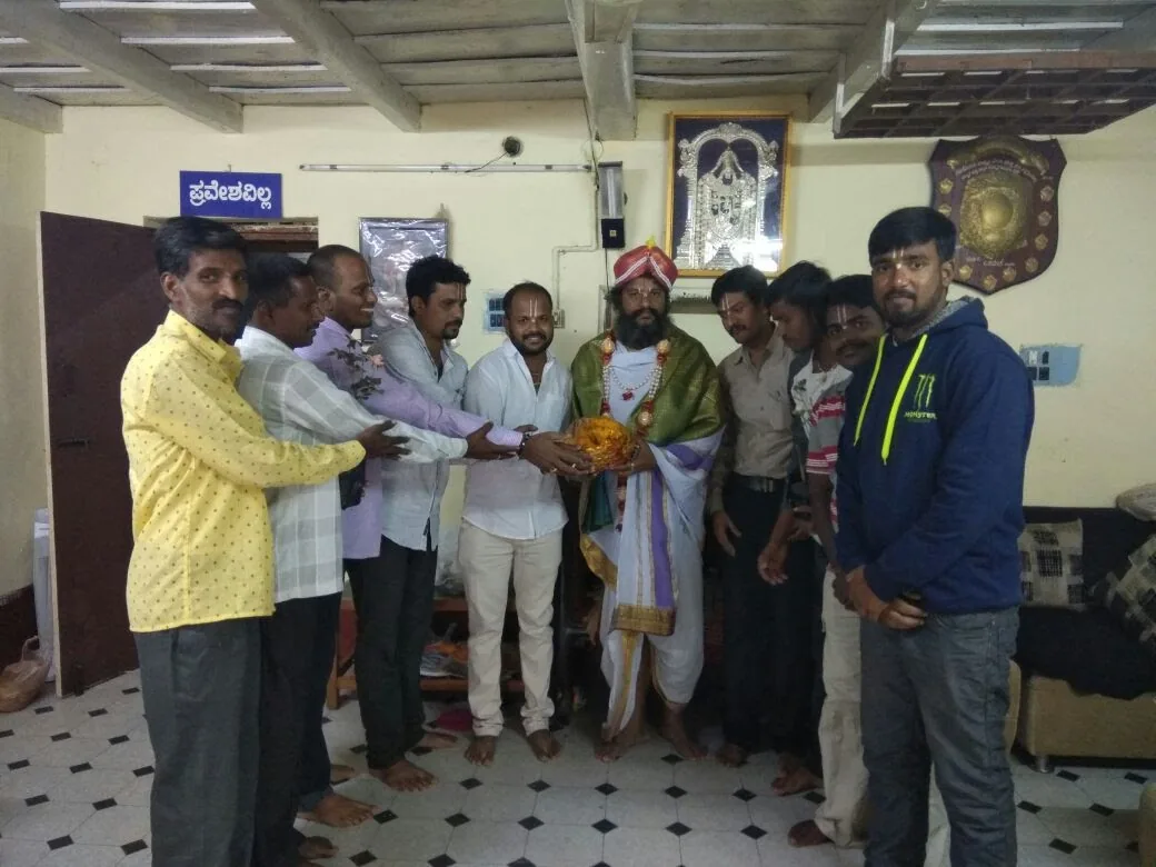 A line of men holding an offering plate out together to Guruji, who stands to receive it in a green shawl and gold turban in the mandira office
