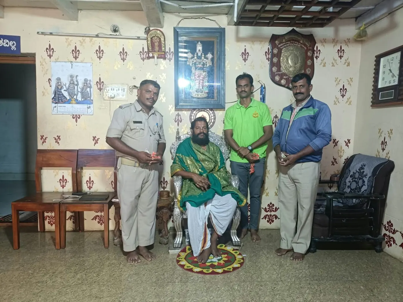 A uniformed police officer and two men standing beside Guruji, who is seated in a green shawl on the silver chair with a rangoli drawn before him