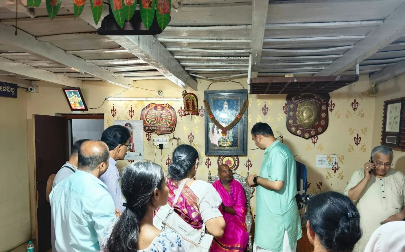 The mandira reception room crowded with visitors standing and talking, one woman being received at the centre and the shrine lit against the far wall
