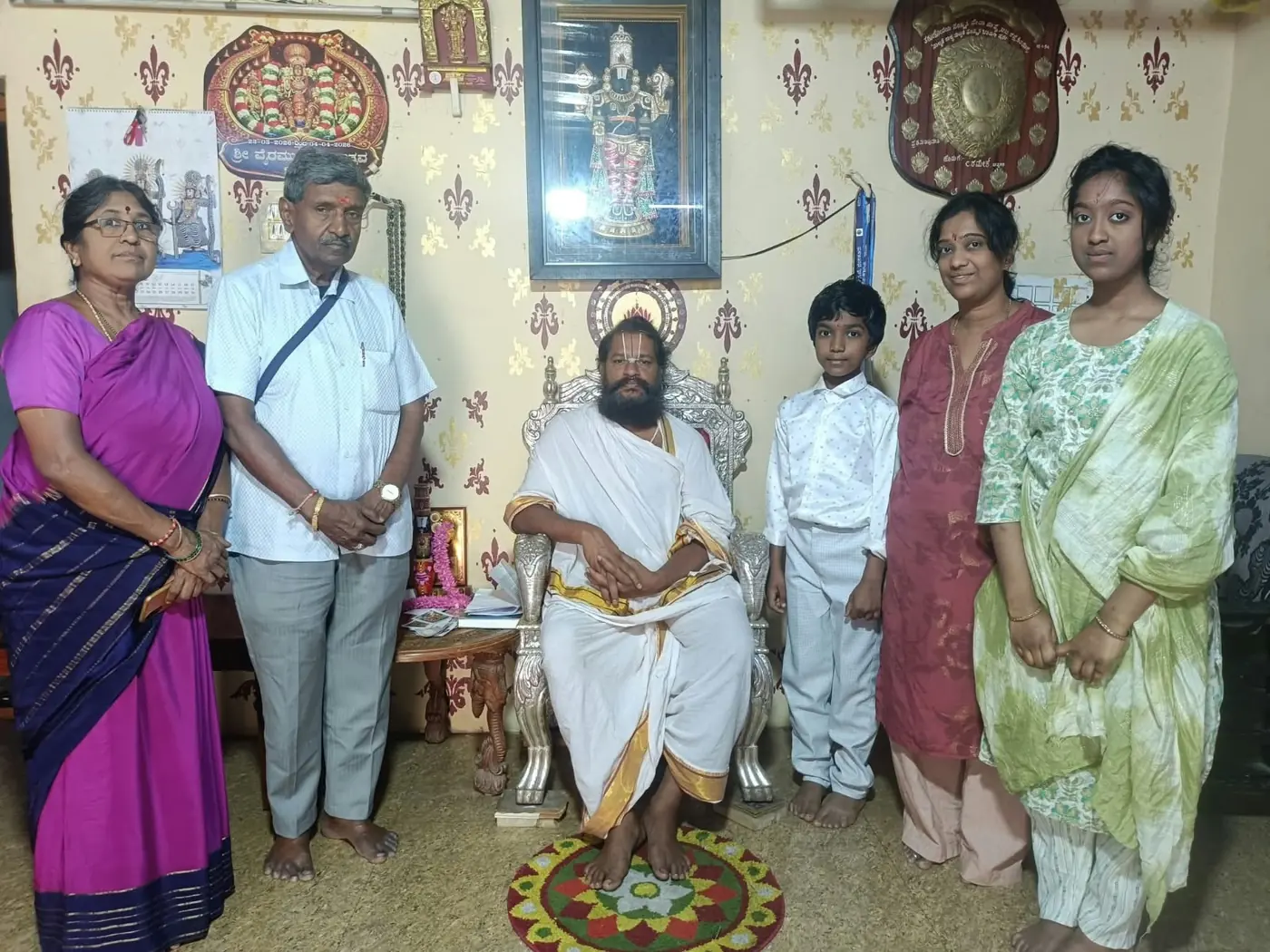 Grandparents, parents and two children standing around Guruji seated in white, the framed deity on the wall above them and a rangoli at his feet