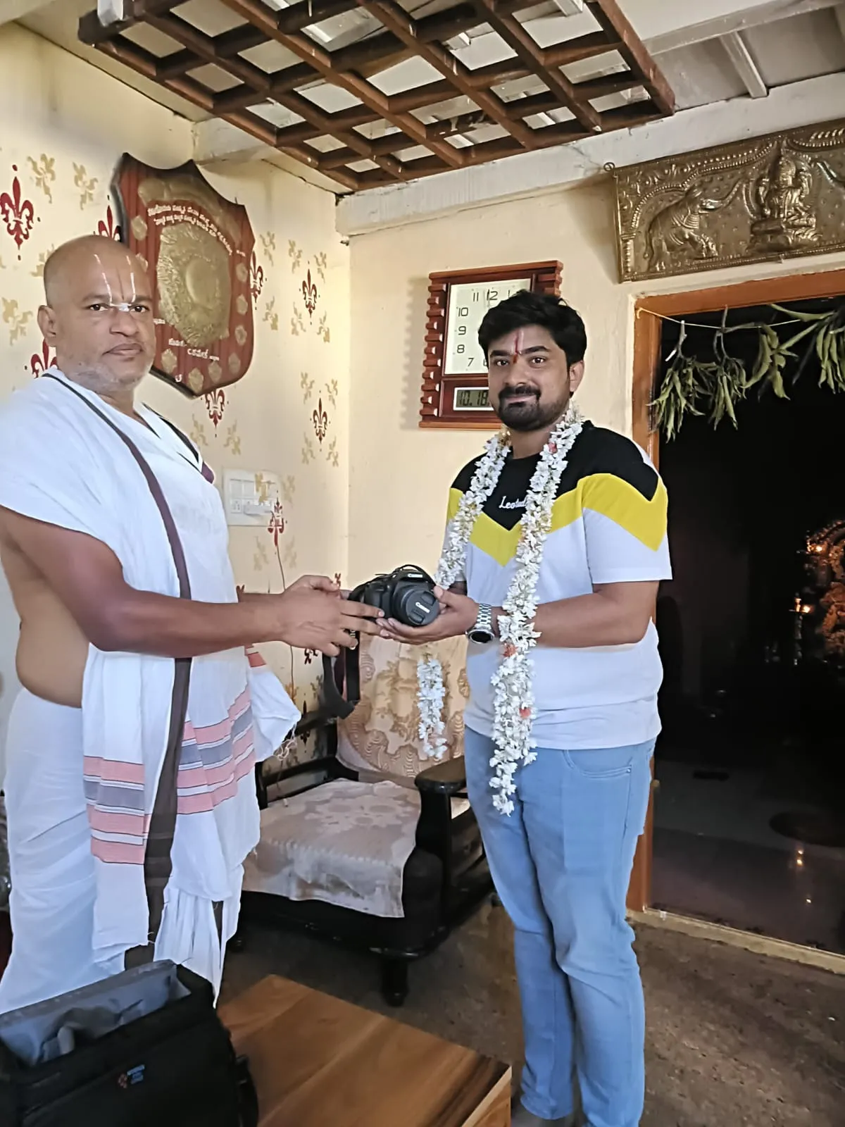 Guruji handing a token of appreciation to a garlanded visitor in a home shrine room