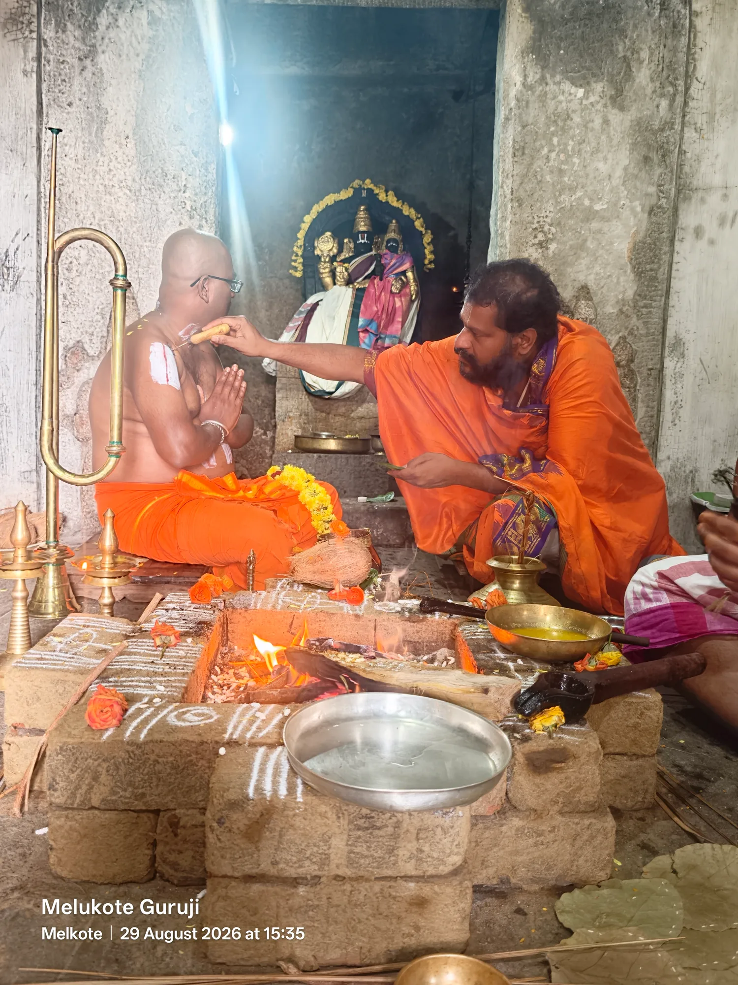 The acharya and a newly initiated disciple in saffron seated on either side of the homa kunda inside a stone shrine, the acharya's hand raised to the disciple's forehead and the deity lit in the niche behind them