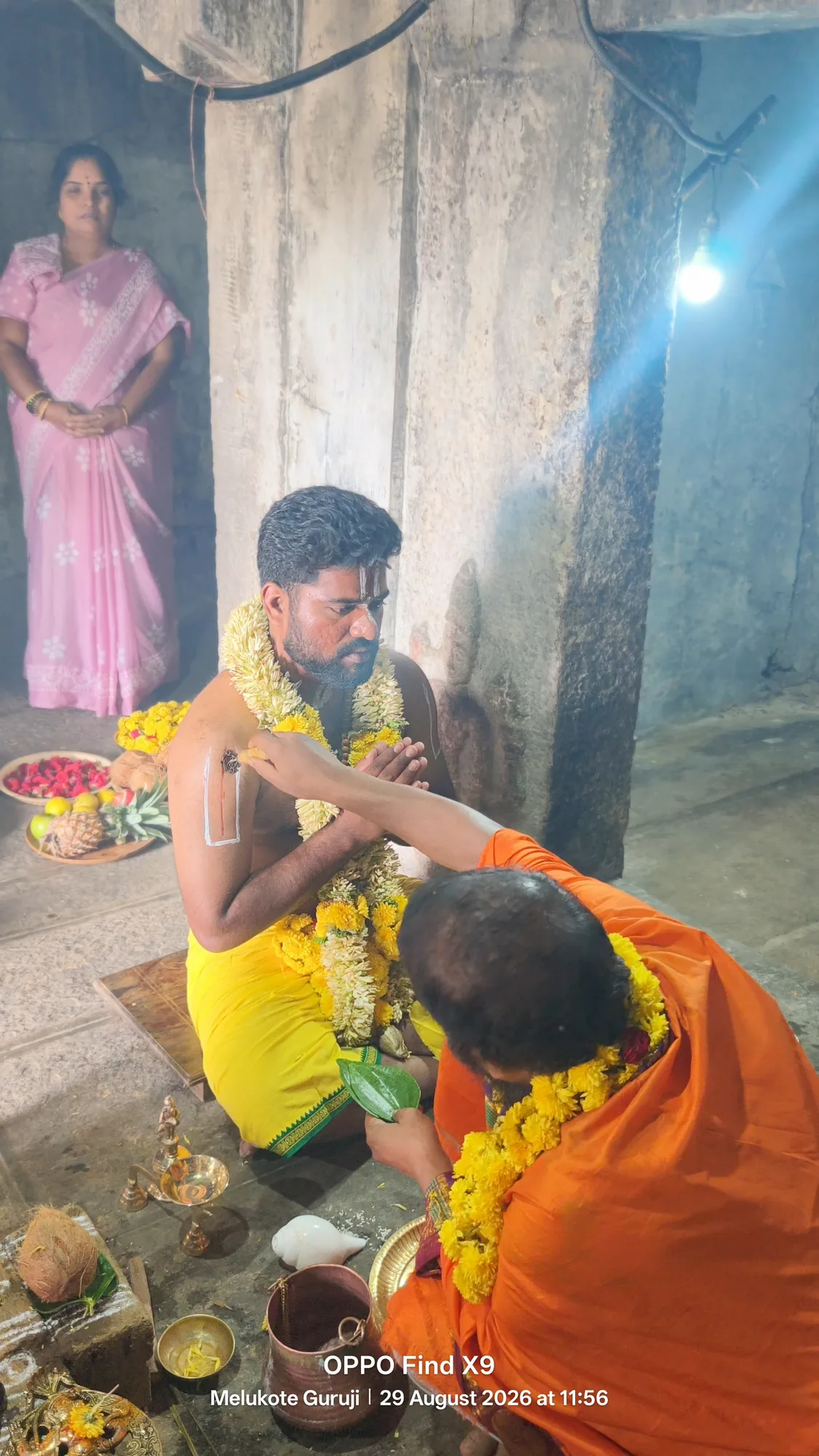 A disciple in a yellow dhoti and heavy garlands sitting with palms joined while the acharya bends to the vessels in front of him, the offerings and the lamp laid out on the stone floor of the shrine