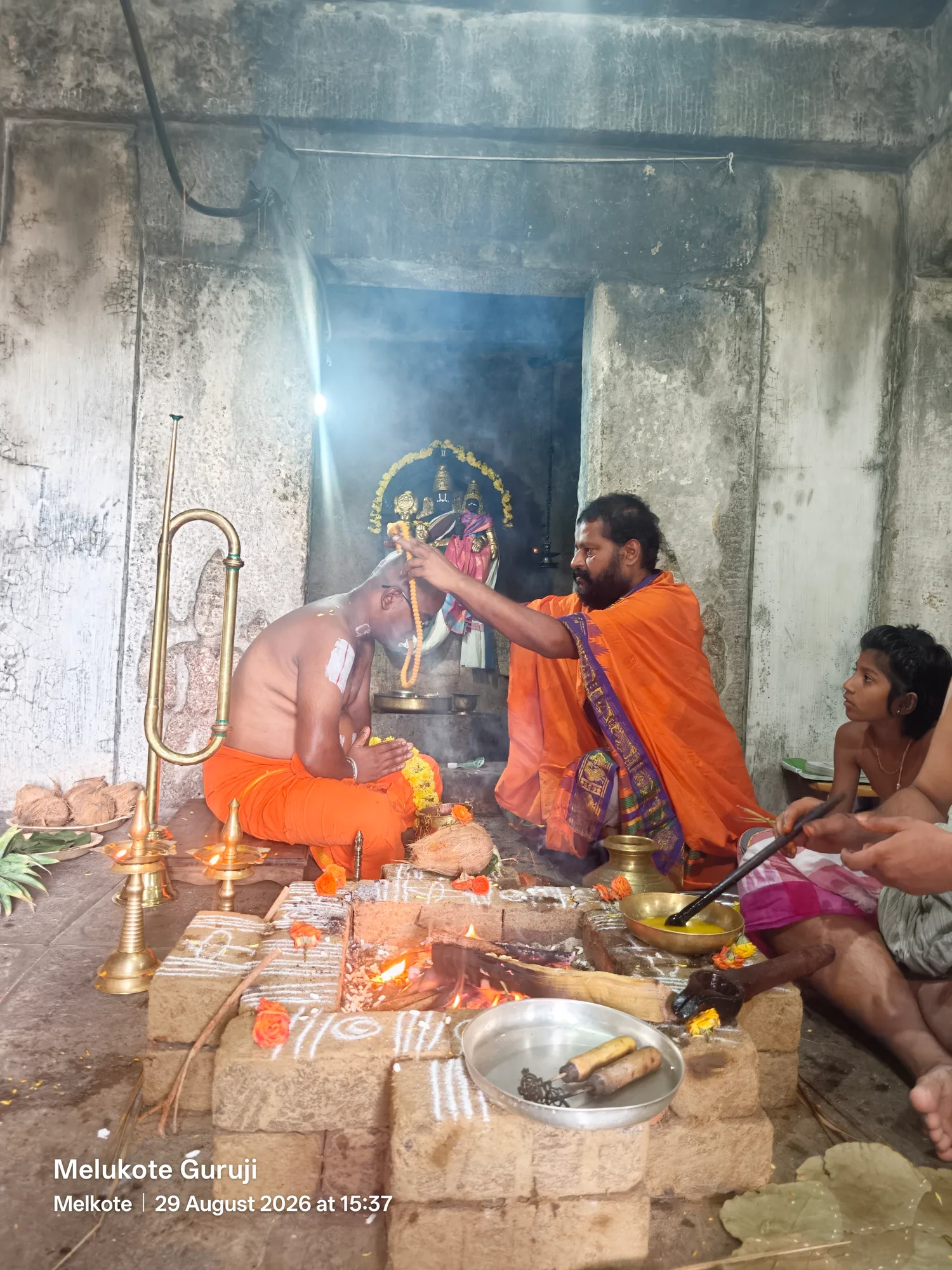 The acharya reaching over the burning homa to lay his hand on the head of the disciple seated opposite him in saffron, a boy watching from the right and the brass staff standing against the shrine wall
