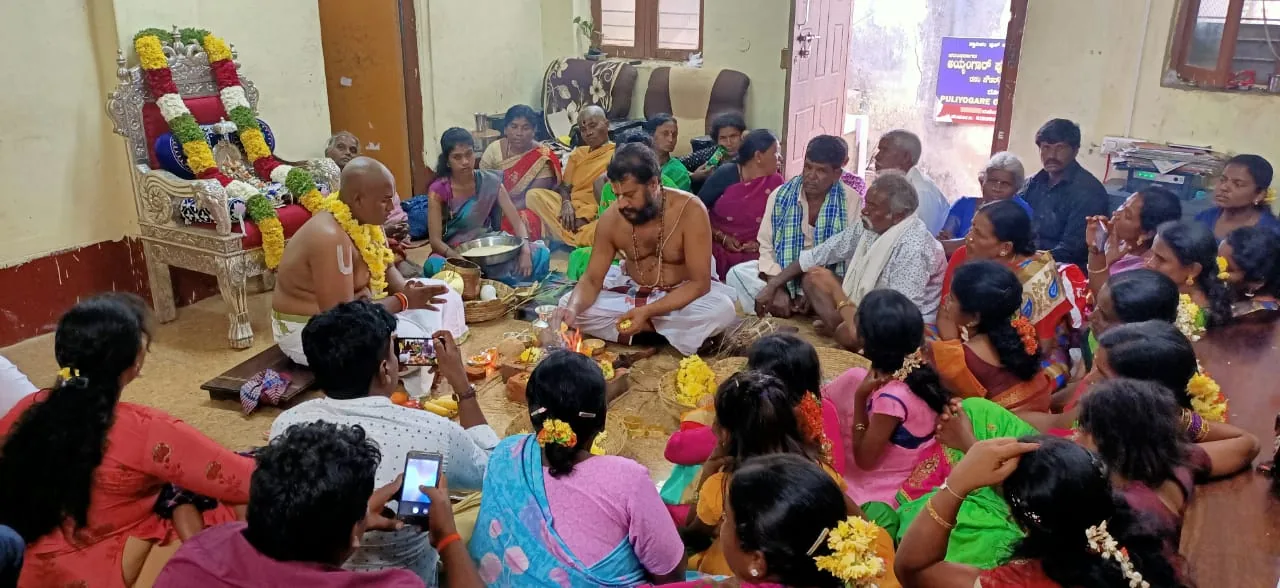 A rite conducted on the floor of a household room packed to the walls with the family watching, the vaidika seated at the offerings with the acharya beside him on a garlanded seat
