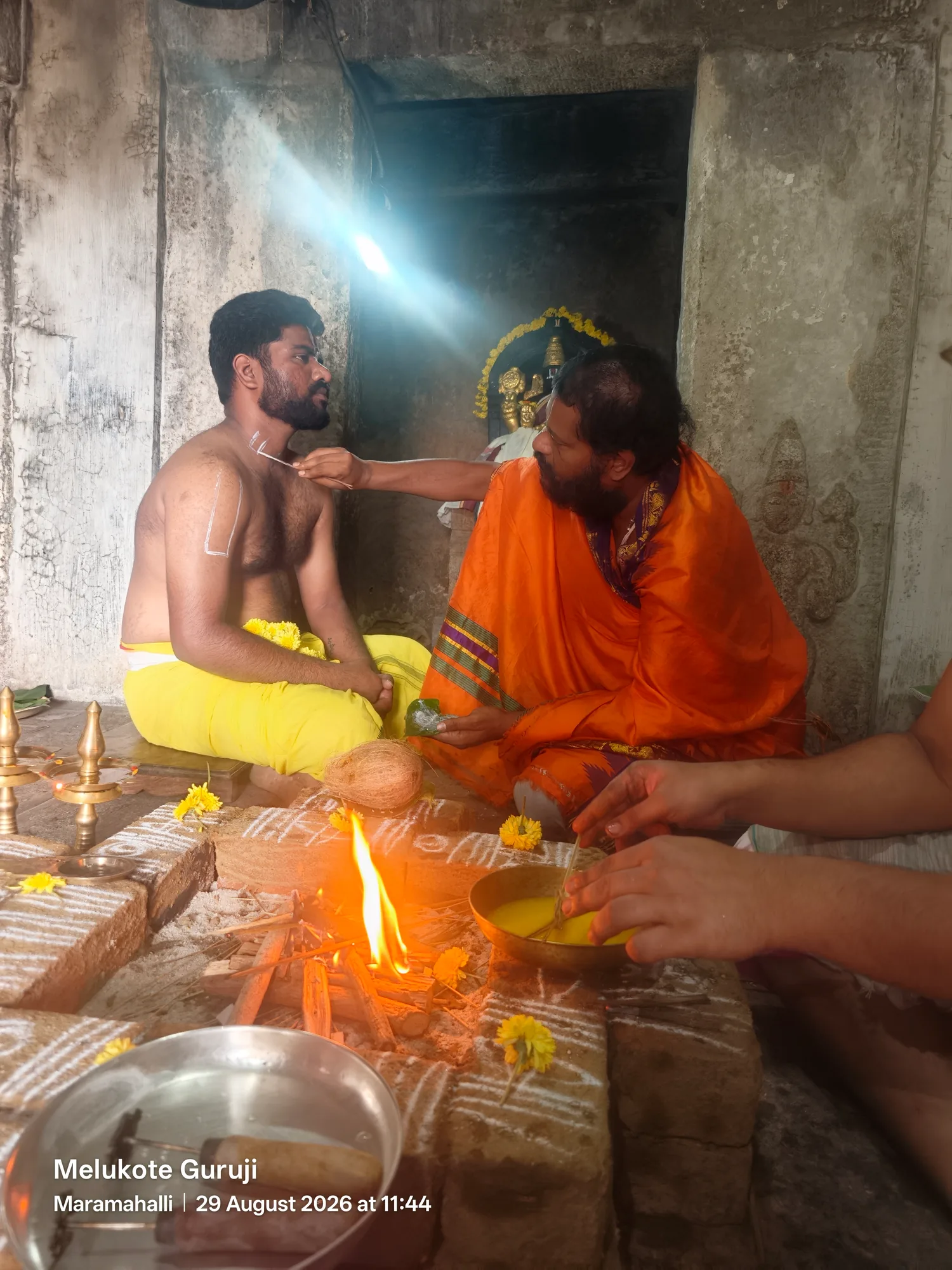 The acharya in saffron leaning across the fire to draw the urdhva pundra on the forehead of a disciple seated bare-shouldered in a yellow dhoti, the homa burning on the brick kunda between them