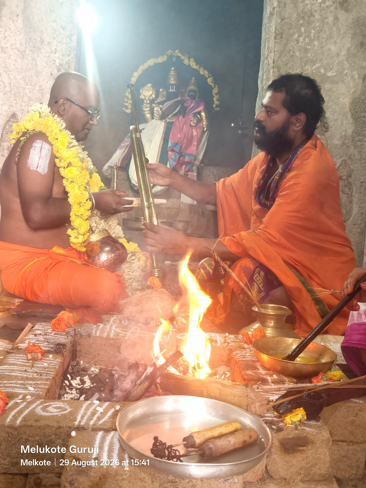 The acharya passing the tridanda staff into the hands of the garlanded disciple across the fire, both of them in saffron, the vessels of the rite ranged along the edge of the kunda