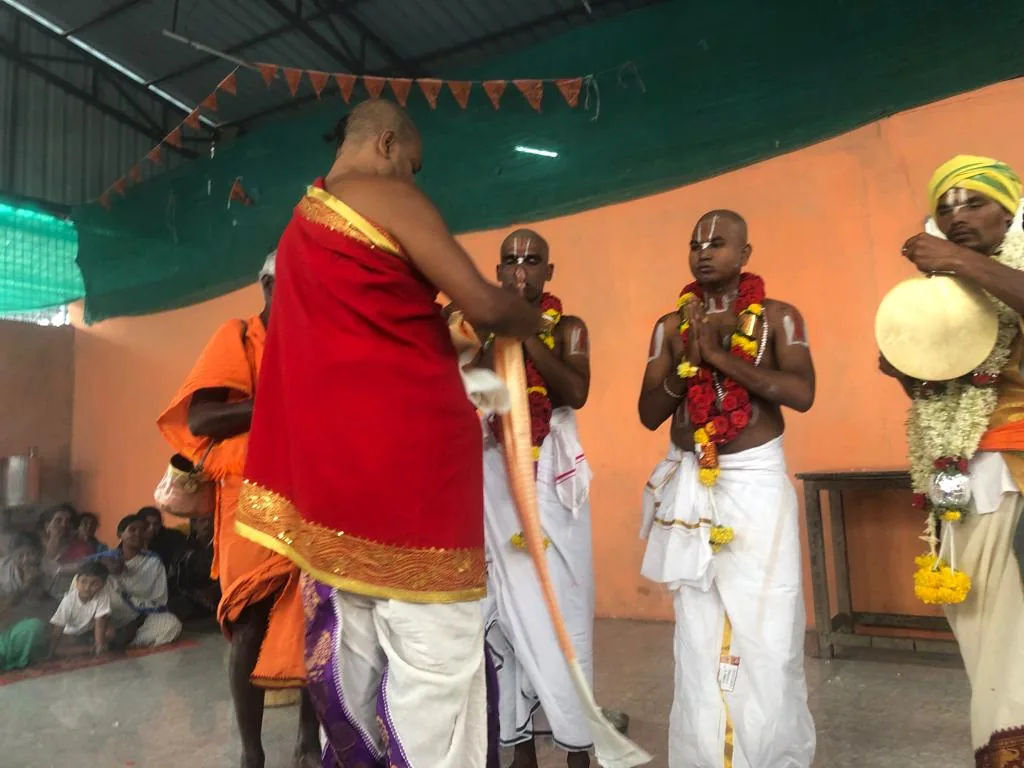 Guruji in a red and gold shawl standing to receive a line of garlanded young disciples in white dhotis, their palms joined, under a hall strung with saffron bunting