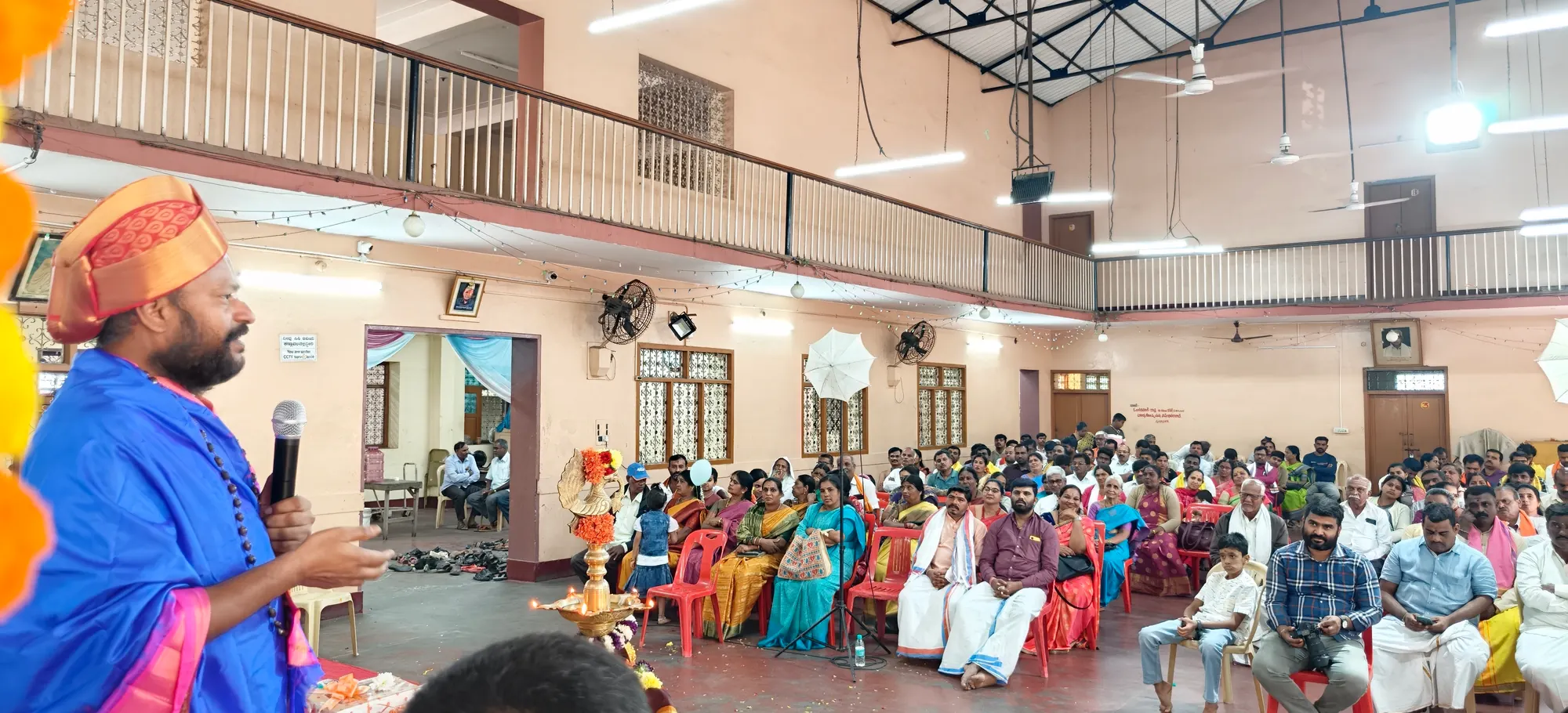 A speaker at the microphone addressing a hall packed with seated listeners, the lamp lit beside him