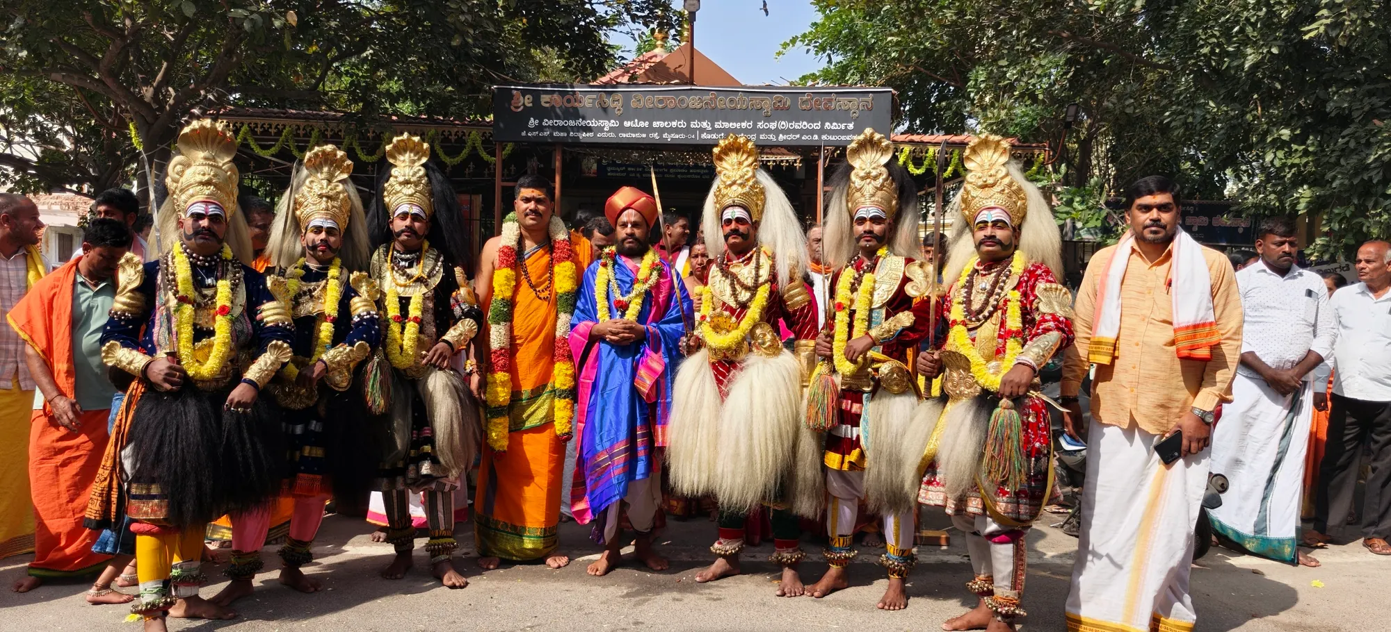 Folk performers in tall crowns and white manes standing in a line outside the gurukulam, garlanded and ready to go on