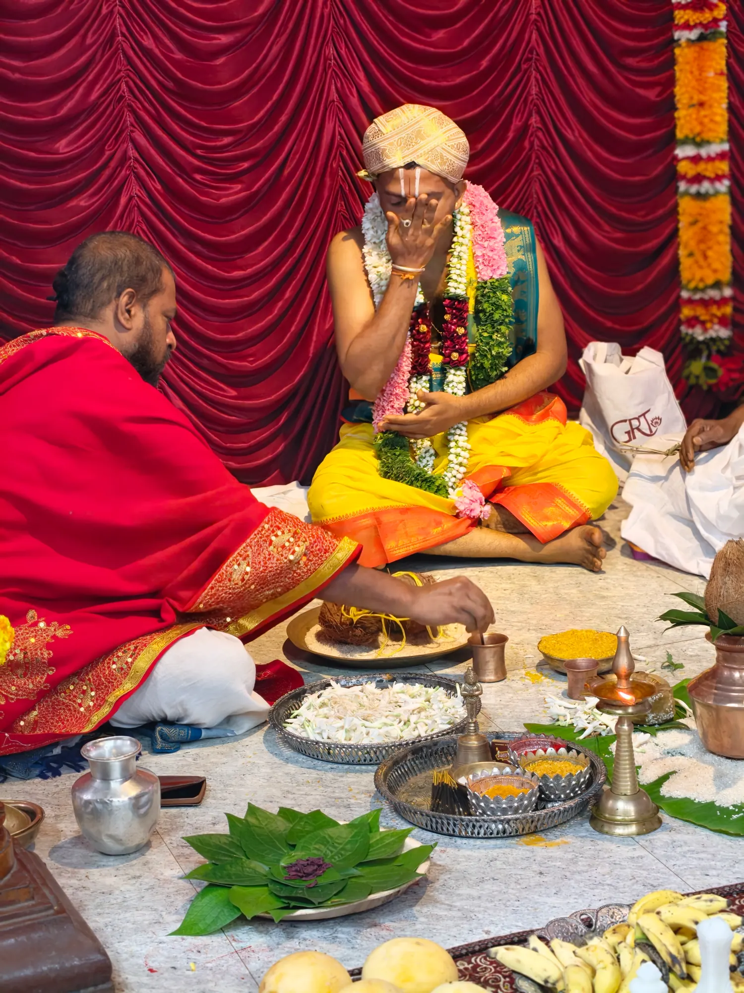 The groom in a gold turban and heavy garlands seated at his part of the rite with his hand raised to his face, the purohita seated opposite him and the vessels laid out between them