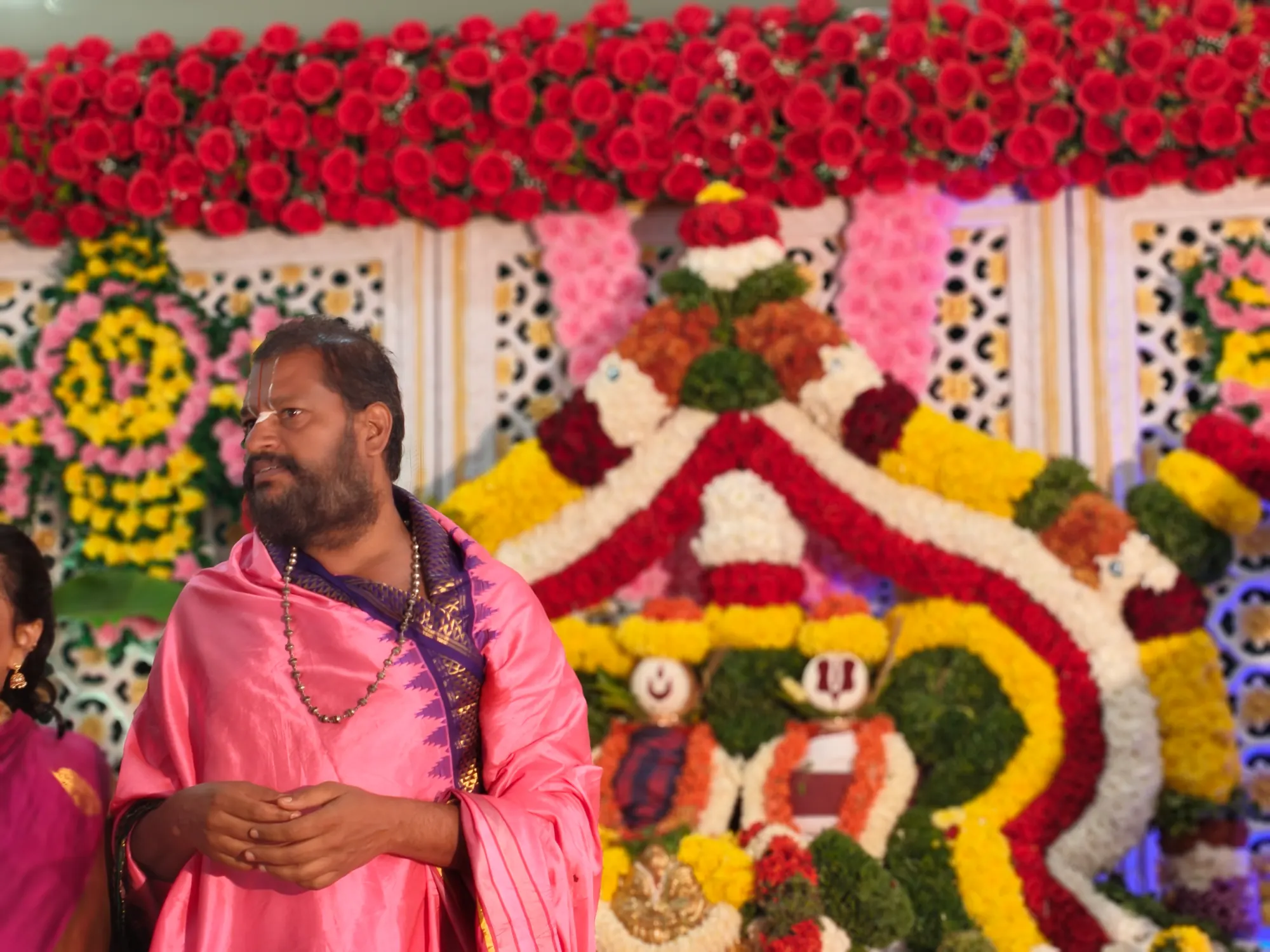 Guruji in a pink shawl standing beside the flower-decorated utsava mandapa, under a canopy of red roses
