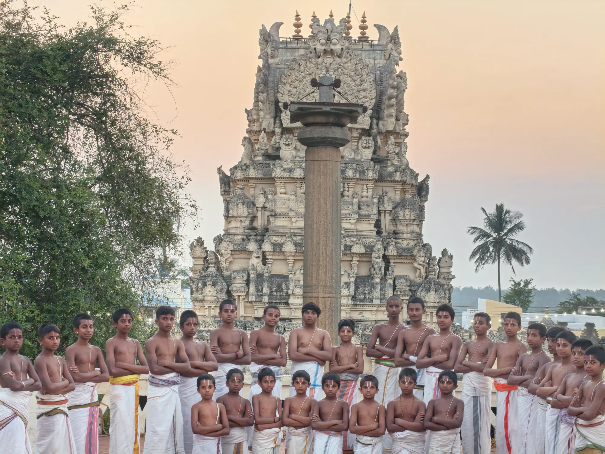 Gurukulam students standing in two rows with their arms folded in front of the temple's Garuda stambha at dusk