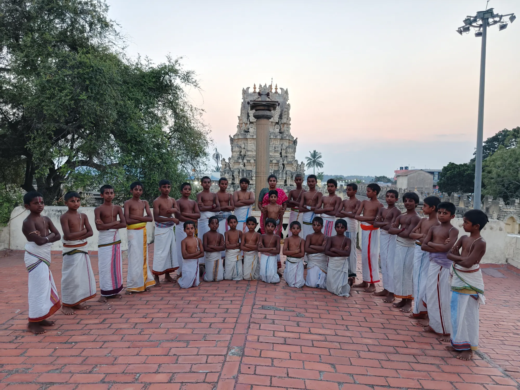 The gurukulam gathered around their teacher on the brick terrace, the Garuda stambha and the gopuram rising behind them at last light