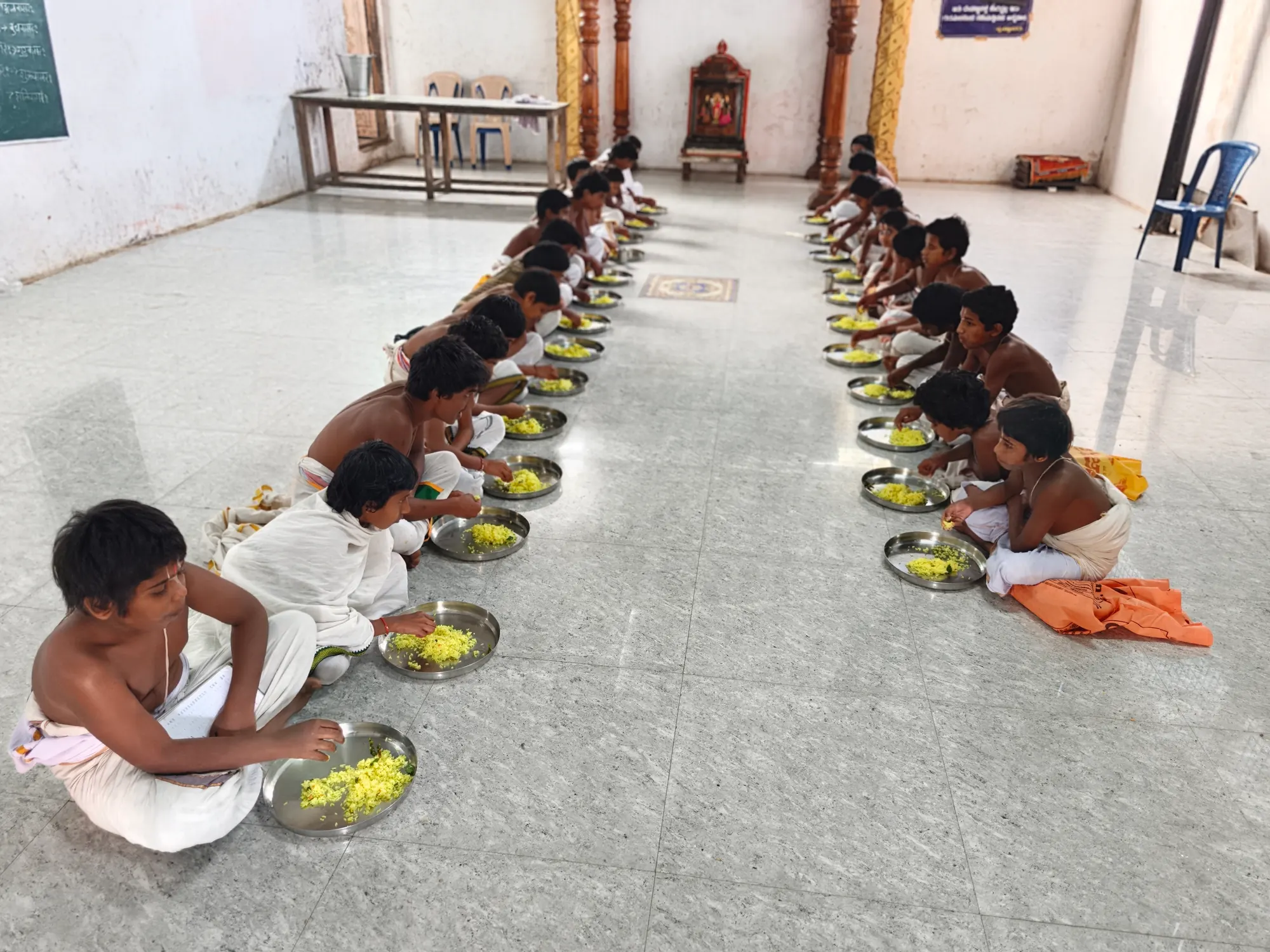 Gurukulam students seated in two long rows down the hall floor, each eating from a steel plate of rice