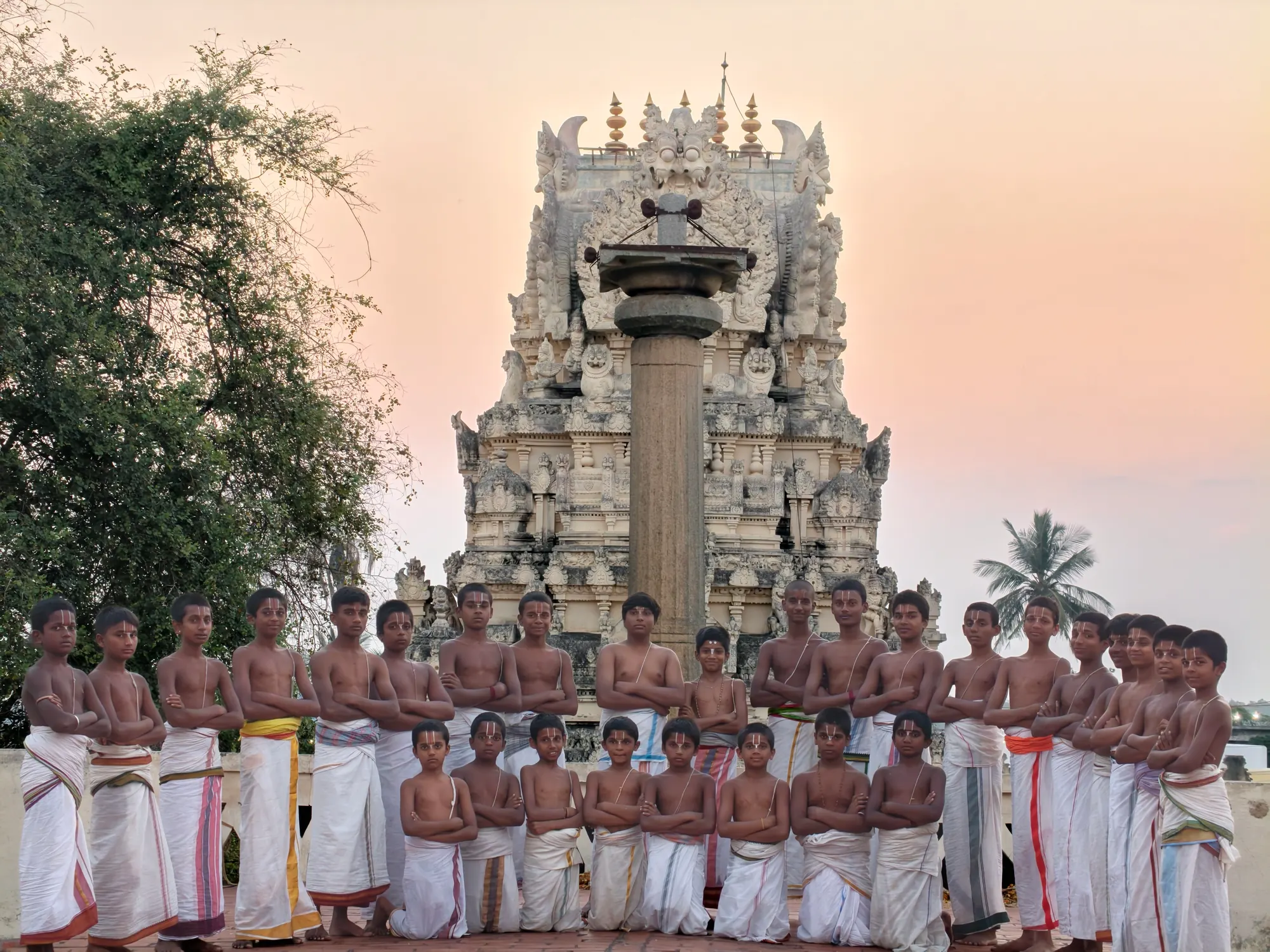 Every student of the gurukulam standing together in white dhotis before the Garuda stambha and the carved gopuram behind it, under a peach sunset sky