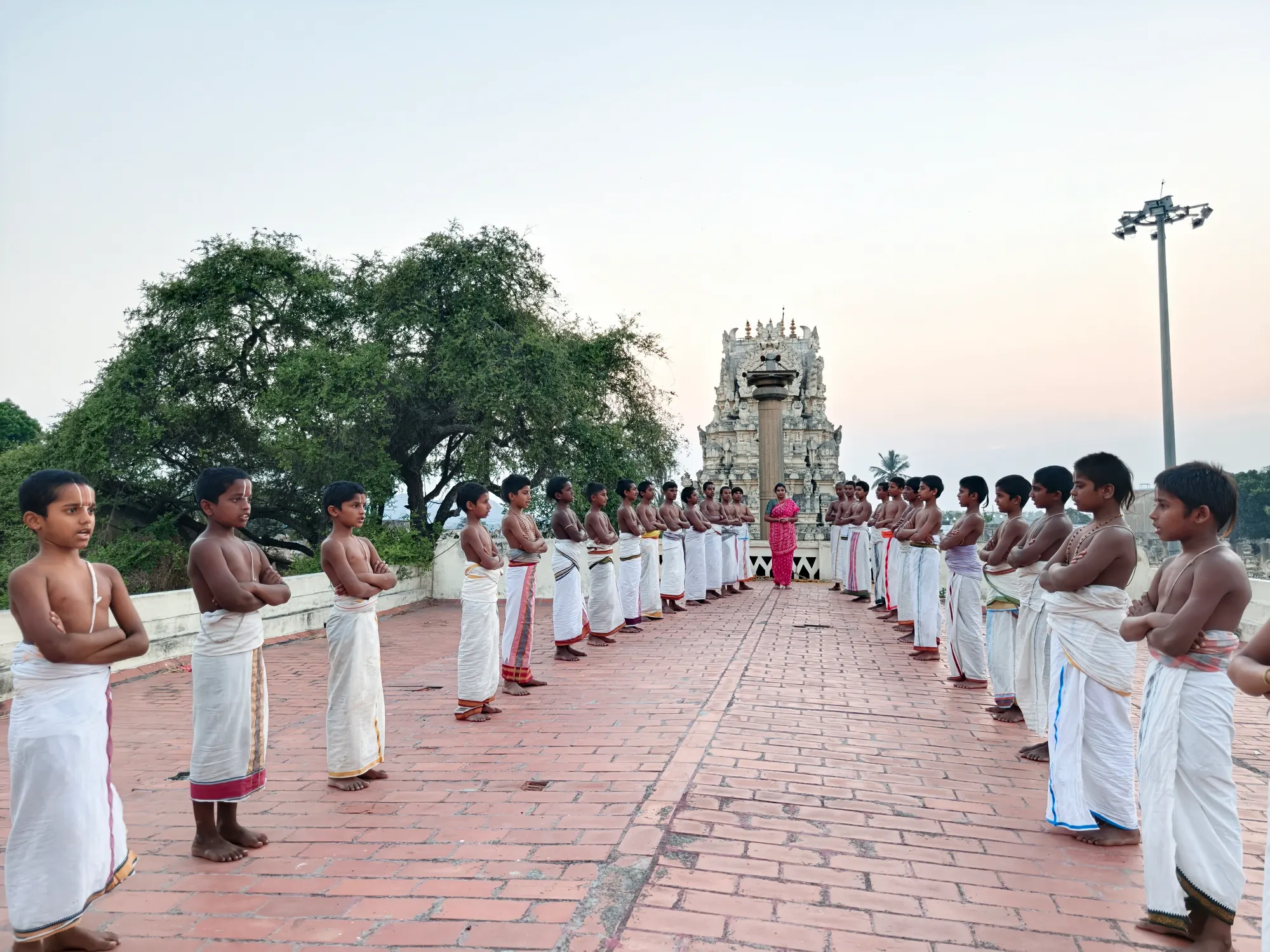 Students in white dhotis standing in two facing lines down the length of the brick terrace, their teacher at the far end before the Garuda stambha