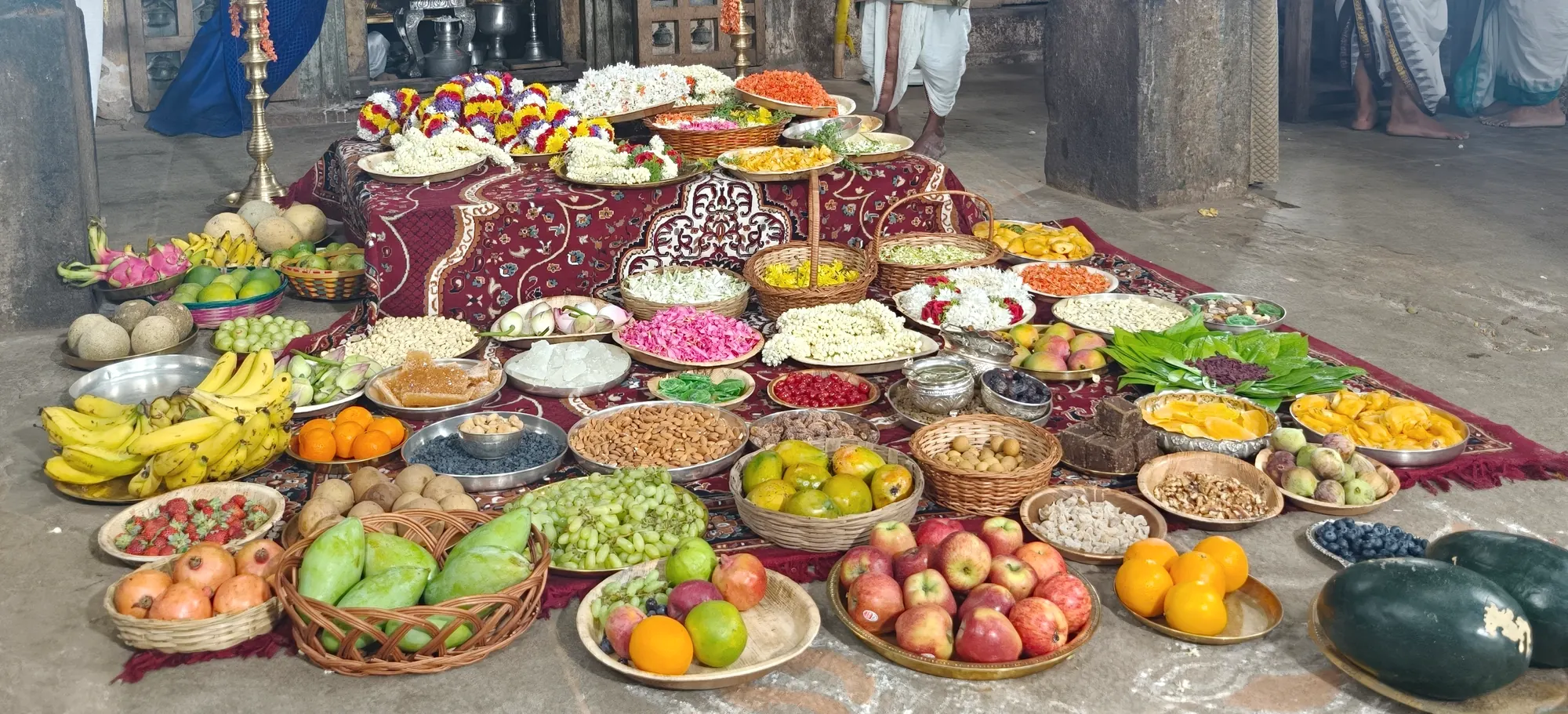 The whole spread of the Hari seve laid out along the floor of the stone hall — plates of fruit, sweets, flowers and grain running the length of the frame
