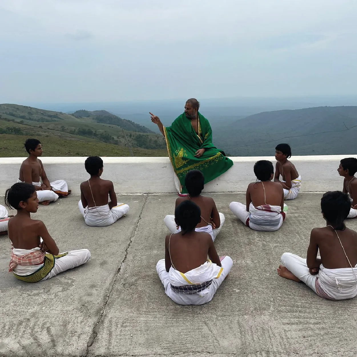 Guruji seated on a parapet teaching a circle of young students, with the hills and valley around Melukote stretching behind them