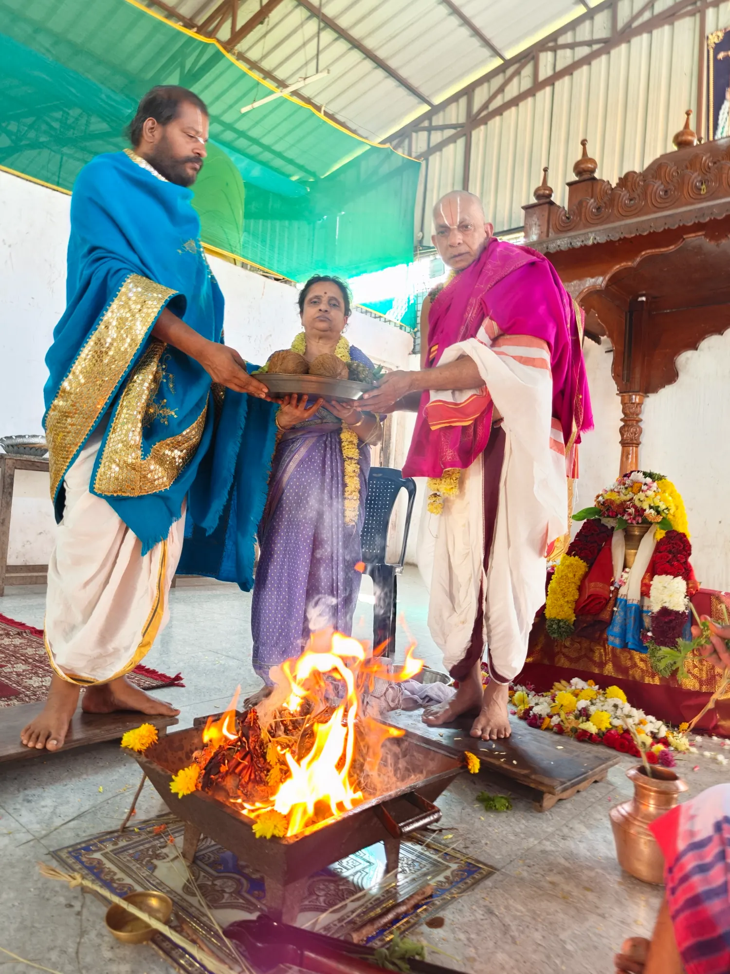 Two priests holding a plate of coconuts together over the homa fire, the woman performing the rite standing between them