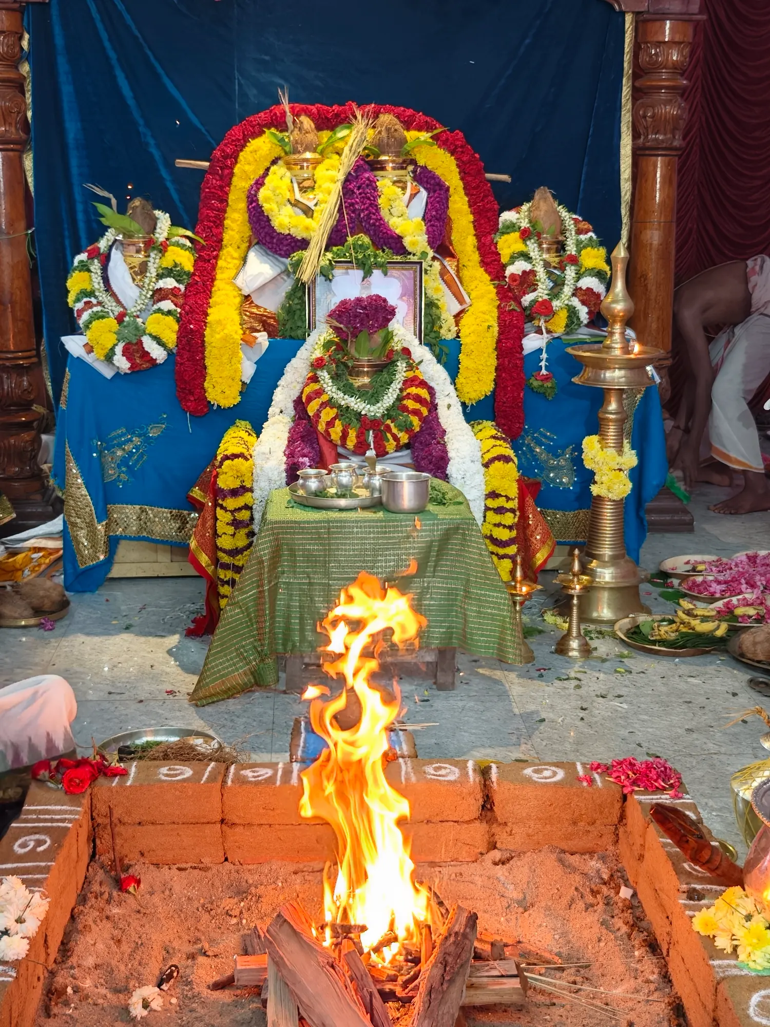 The homa fire burning high in its brick kunda before a garlanded altar of kalashas, a framed image and a tall brass lamp