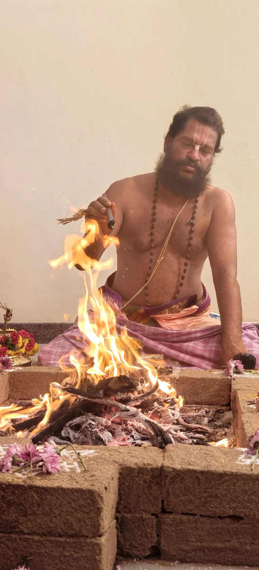 A vaidika seated at the brick homa kunda holding a lit torch of grass down to the fire, the flame rising between him and the camera
