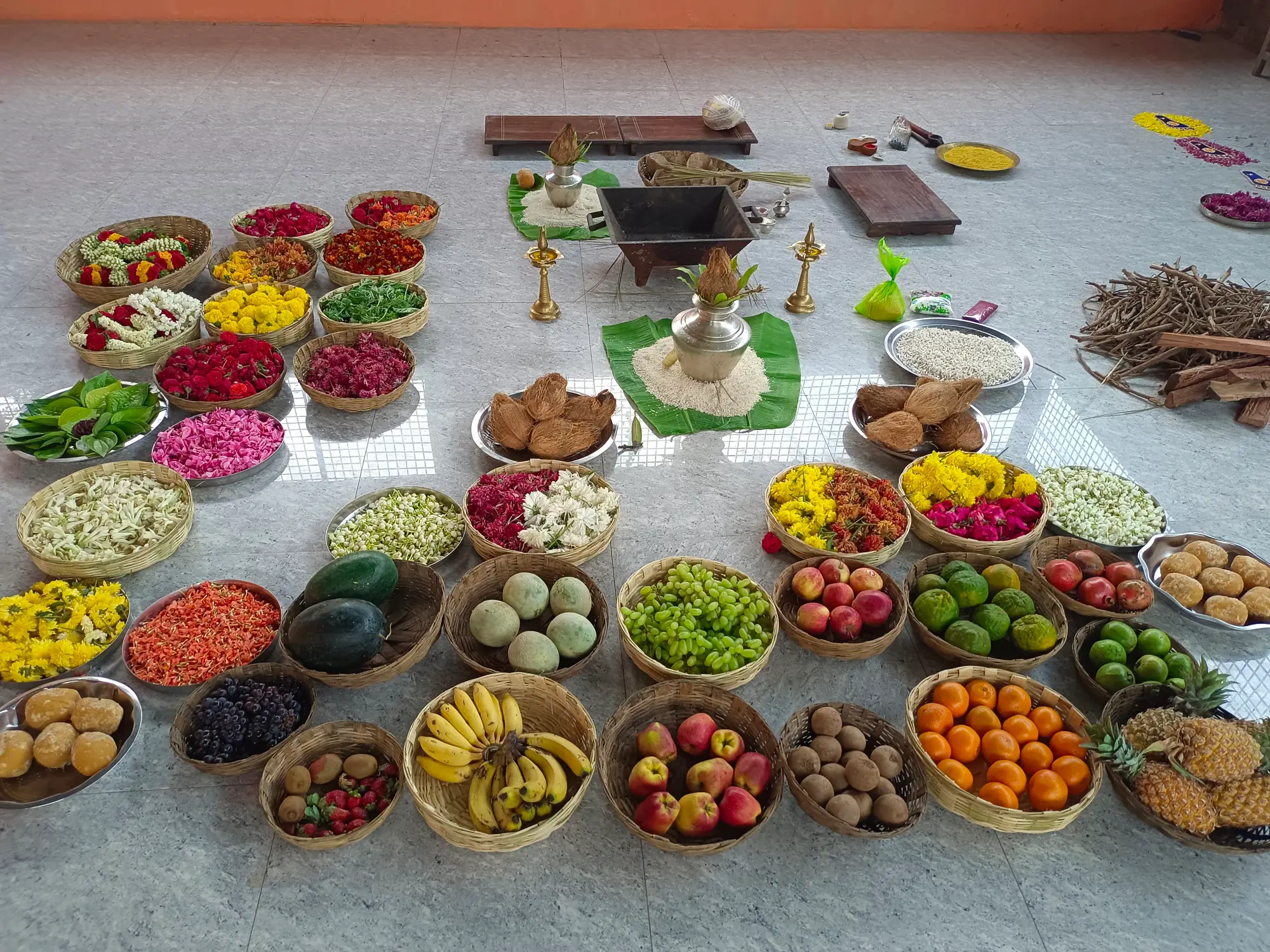 The offerings laid out on the floor before a homa begins — baskets of fruit and flowers, mounds of grain, the kalasha on a banana leaf, and firewood stacked at the edge